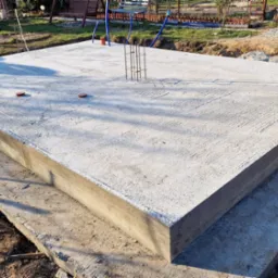 Wylana płyta fundamentowa z wystającymi prętami zbrojeniowymi, widoczna struktura betonu i ślady szalunku. Tło: trawa, ziemia, elementy konstrukcji. Ujęcie z niskiej perspektywy.