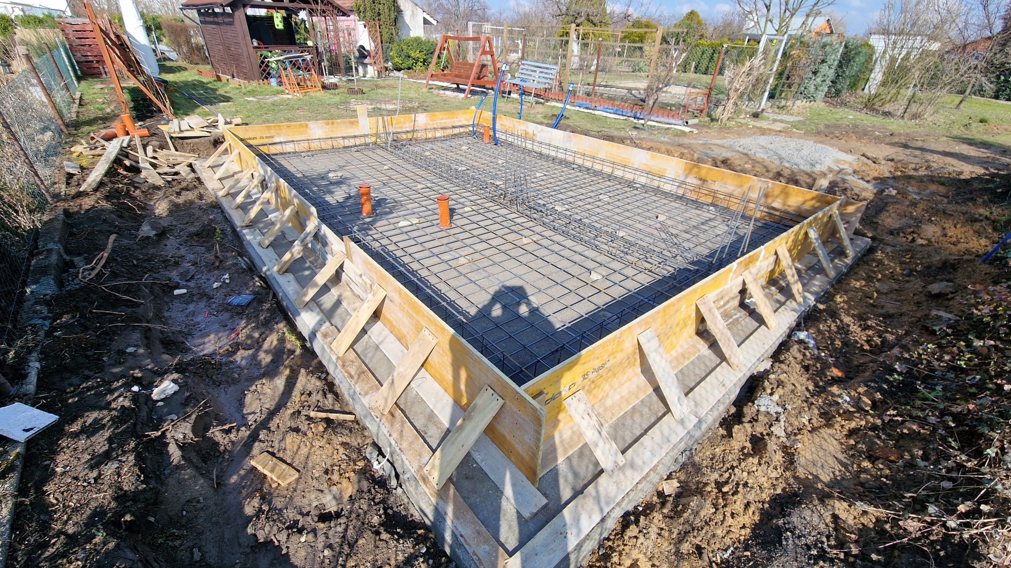Przygotowany szalunek i zbrojenie pod płytę fundamentową na działce. Widoczne pomarańczowe rury instalacyjne. Ziemia wokół wykopu.