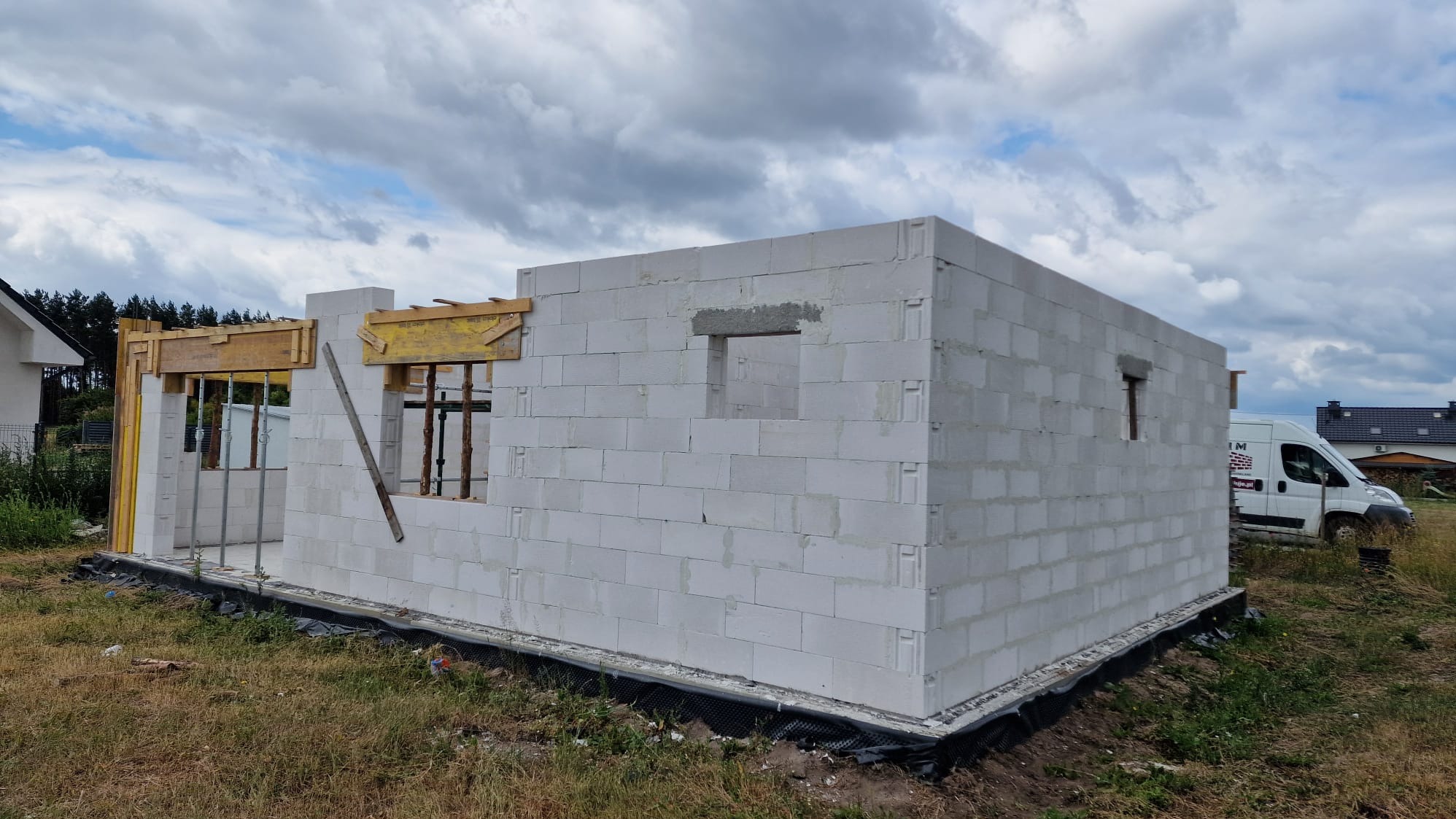 Wznoszenie ścian z betonu komórkowego; widoczne drewniane szalunki okienne i drzwiowe. Na drugim planie budynek mieszkalny i samochód dostawczy.