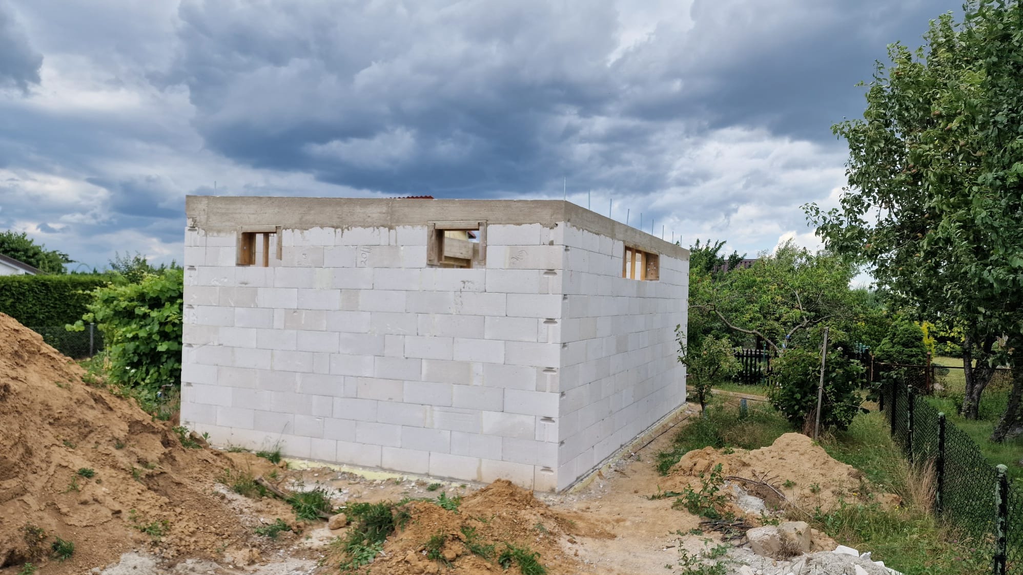 Niski budynek z białych bloczków, z otworami okiennymi z drewnianymi ramami, na fundamencie, w otoczeniu zieleni i pod pochmurnym niebem.