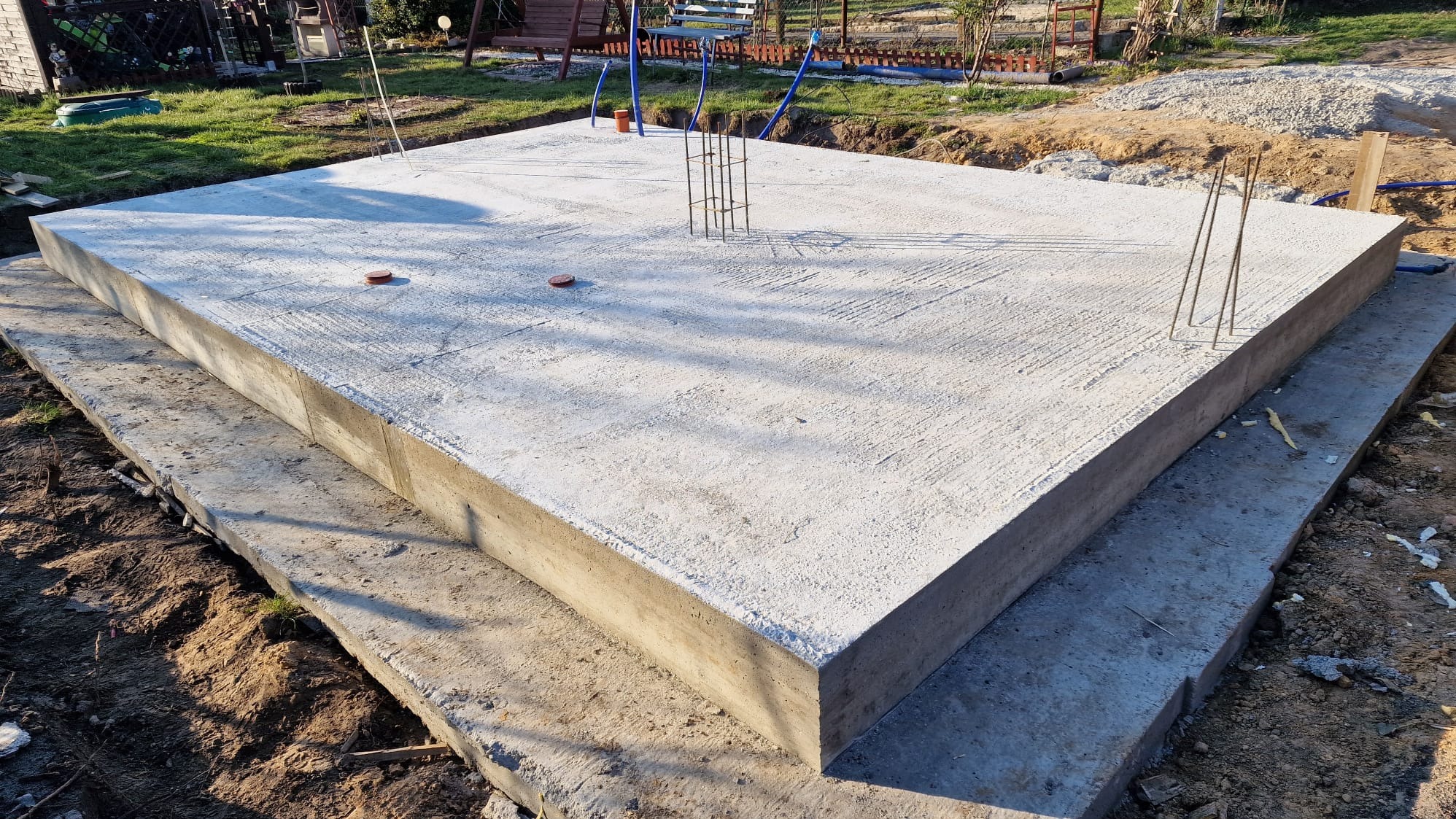 Wylana płyta fundamentowa z wystającymi prętami zbrojeniowymi, widoczna struktura betonu i ślady szalunku. Tło: trawa, ziemia, elementy konstrukcji. Ujęcie z niskiej perspektywy.