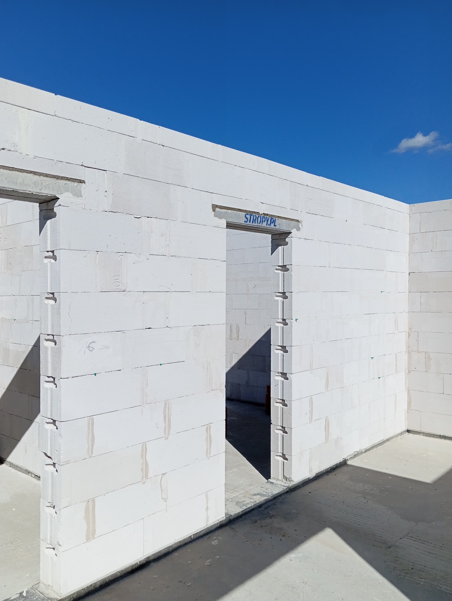 Wnętrze domu w trakcie budowy z białych bloczków, widoczne otwory drzwiowe i niebieski napis 'STROPY.PL' nad jednym z nich, błękitne niebo w tle.