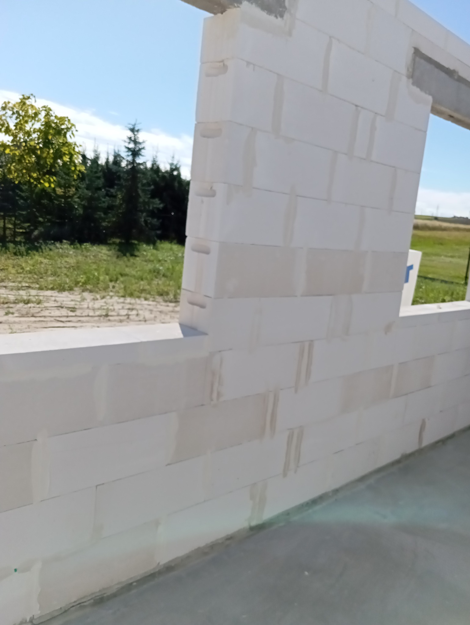 Ściana z białych bloczków, widoczne spoiny. W tle zieleń drzew i błękit nieba. Budowa domu w trakcie wznoszenia ścian nośnych. Widoczny fragment otworu okiennego.