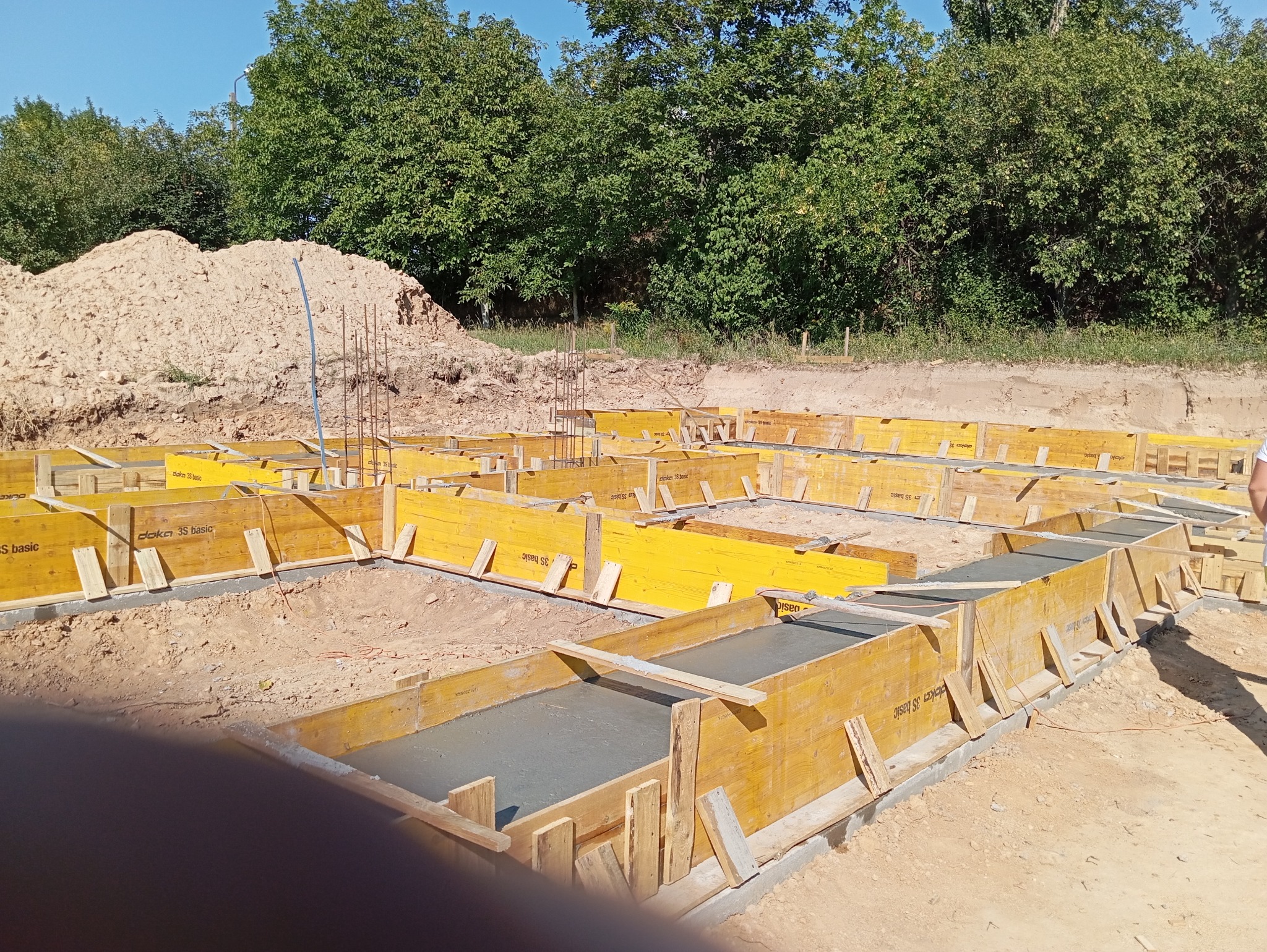 Żółte szalunki fundamentów wypełnione świeżym betonem, widok z góry na plac budowy z hałdą ziemi w tle i zielenią drzew. Szalunki marki Doka 3S basic.