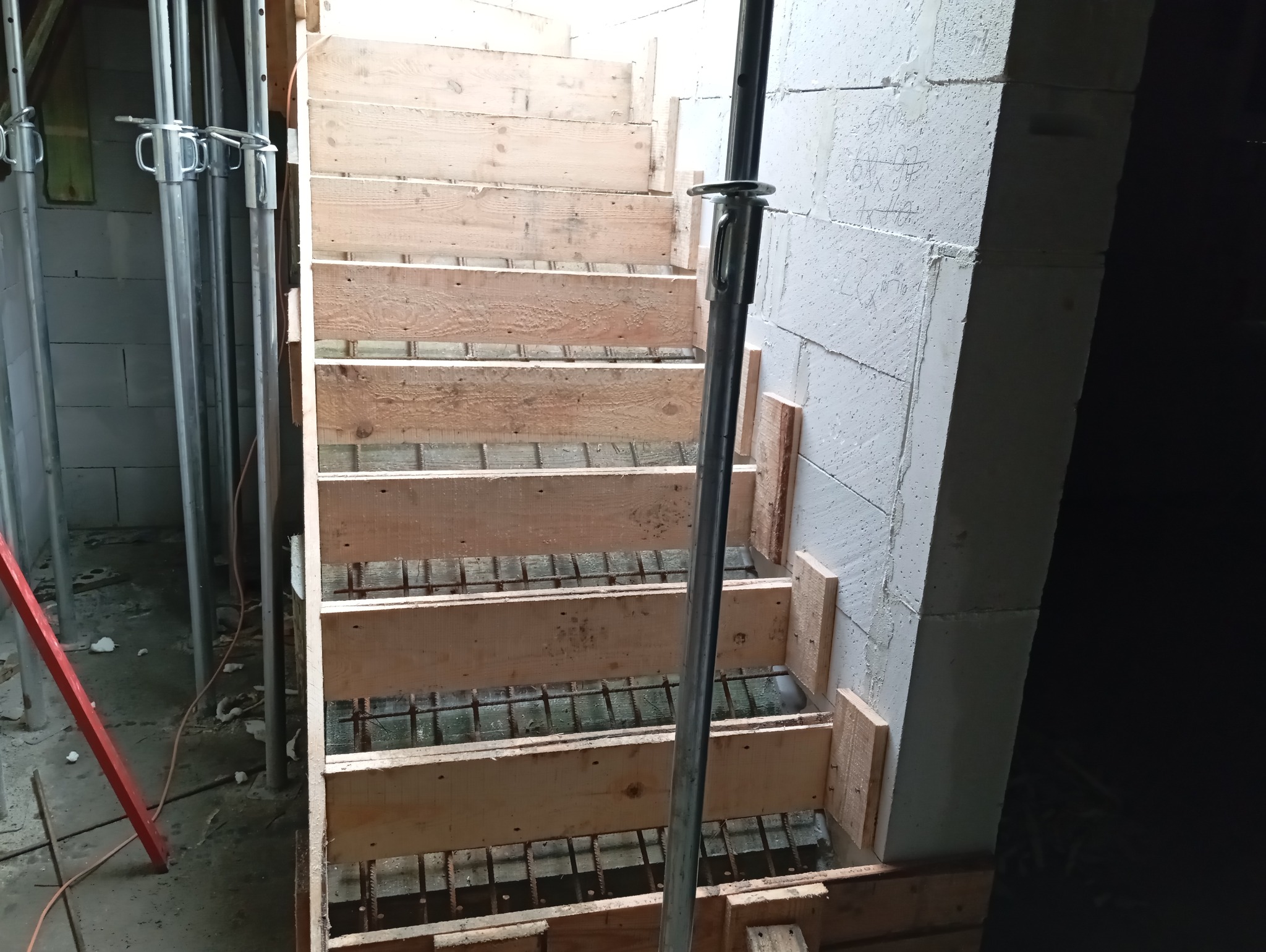 Szczegółowe ujęcie drewnianego szalunku schodów z widocznym zbrojeniem, wspartego metalowymi stemplami budowlanymi, przy ścianie z betonu komórkowego.