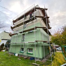 Sunclean - mycie elewacji, dachów, paneli fotowoltaicznych - Dom w rusztowaniach, przygotowywany do prac elewacyjnych. Widoczny fragment elewacji w trakcie czyszczenia. Zielona trawa i drzewa w tle.