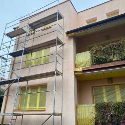 Sunclean - mycie elewacji, dachów, paneli fotowoltaicznych - Budynek w trakcie zabezpieczania elewacji folią i taśmą malarską przed pracami. Widoczne rusztowania i kwiaty na balkonie. Ujęcie z dołu.