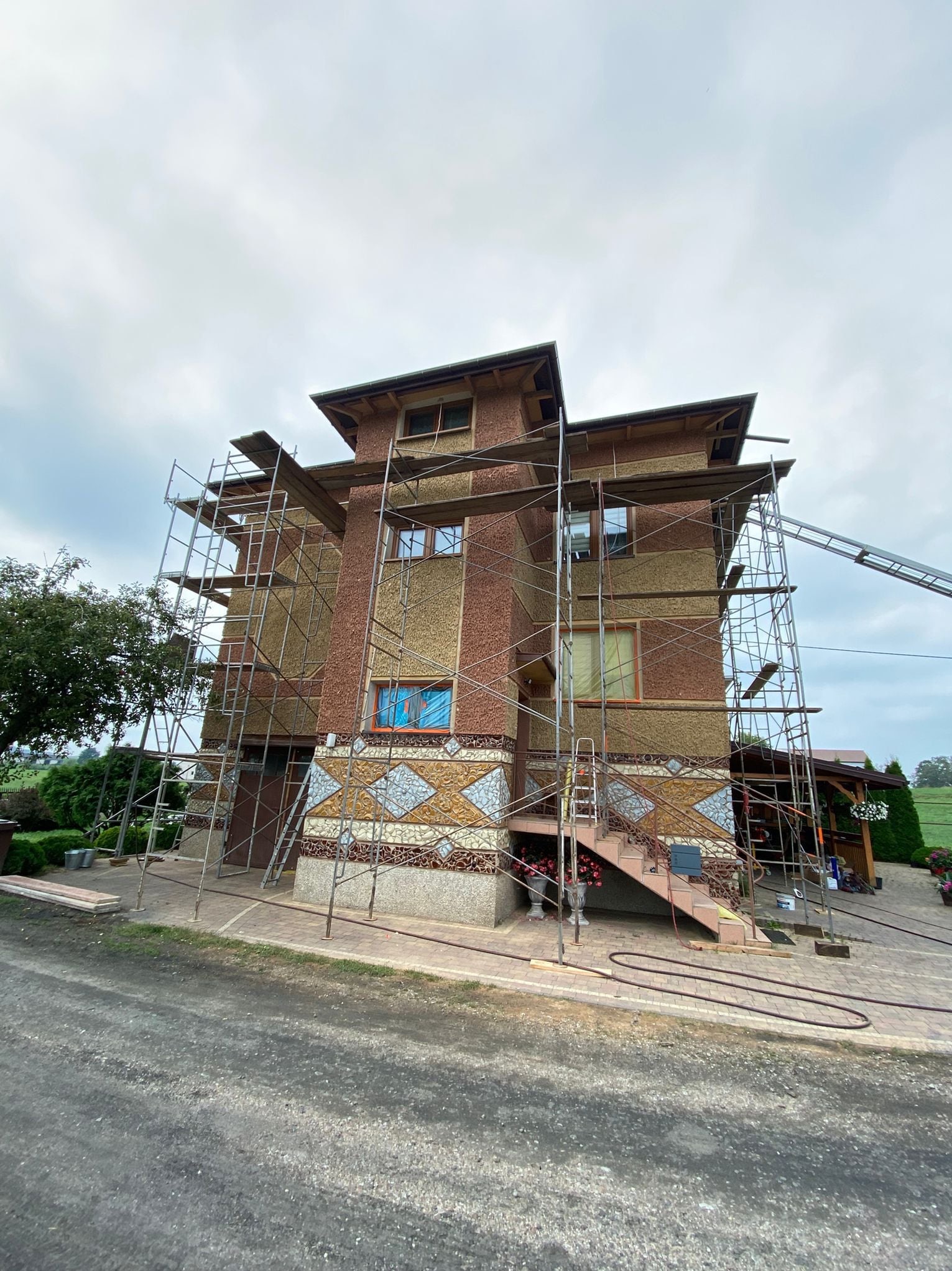 Dom w trakcie prac elewacyjnych, z widocznymi rusztowaniami. Elewacja z dekoracyjnymi elementami, przygotowana do malowania. Widok z dołu, pochmurne niebo.