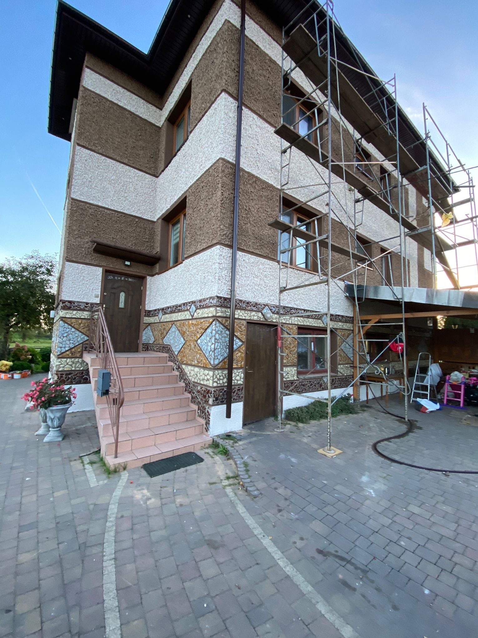 Dwupiętrowy dom po malowaniu elewacji, z dekoracyjnym wykończeniem mozaikowym u podstawy. Widoczne rusztowania i schody wejściowe z donicami kwiatów.