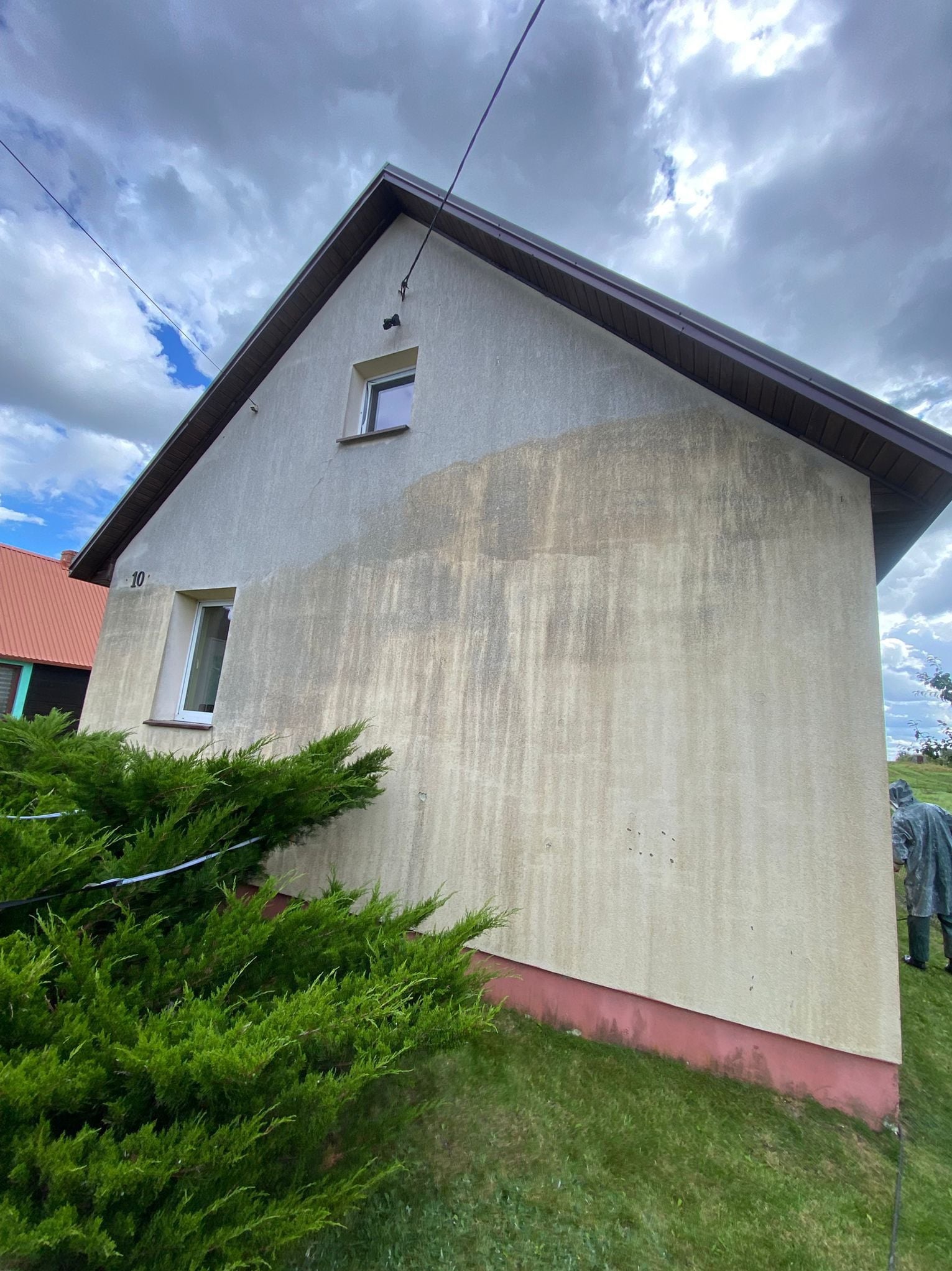 Fragment ściany domu z numerem 10, w trakcie czyszczenia. Widoczna różnica w kolorze elewacji przed i po umyciu. Częściowo zasłonięta przez krzew.