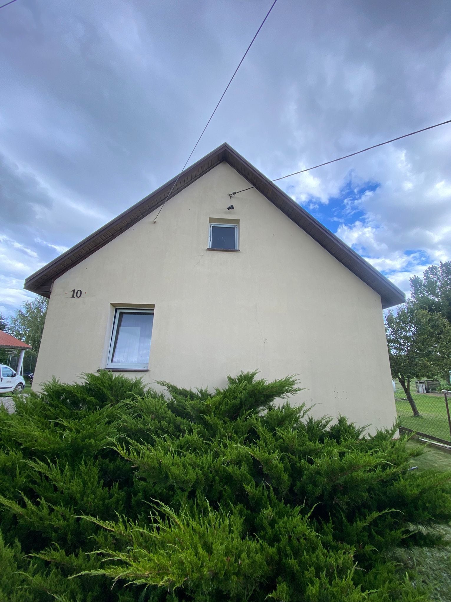 Jasna elewacja domu jednorodzinnego z numerem 10, widoczna z dołu, po zabiegu czyszczenia. Krzewy iglaste w pierwszym planie, błękitne niebo w tle.