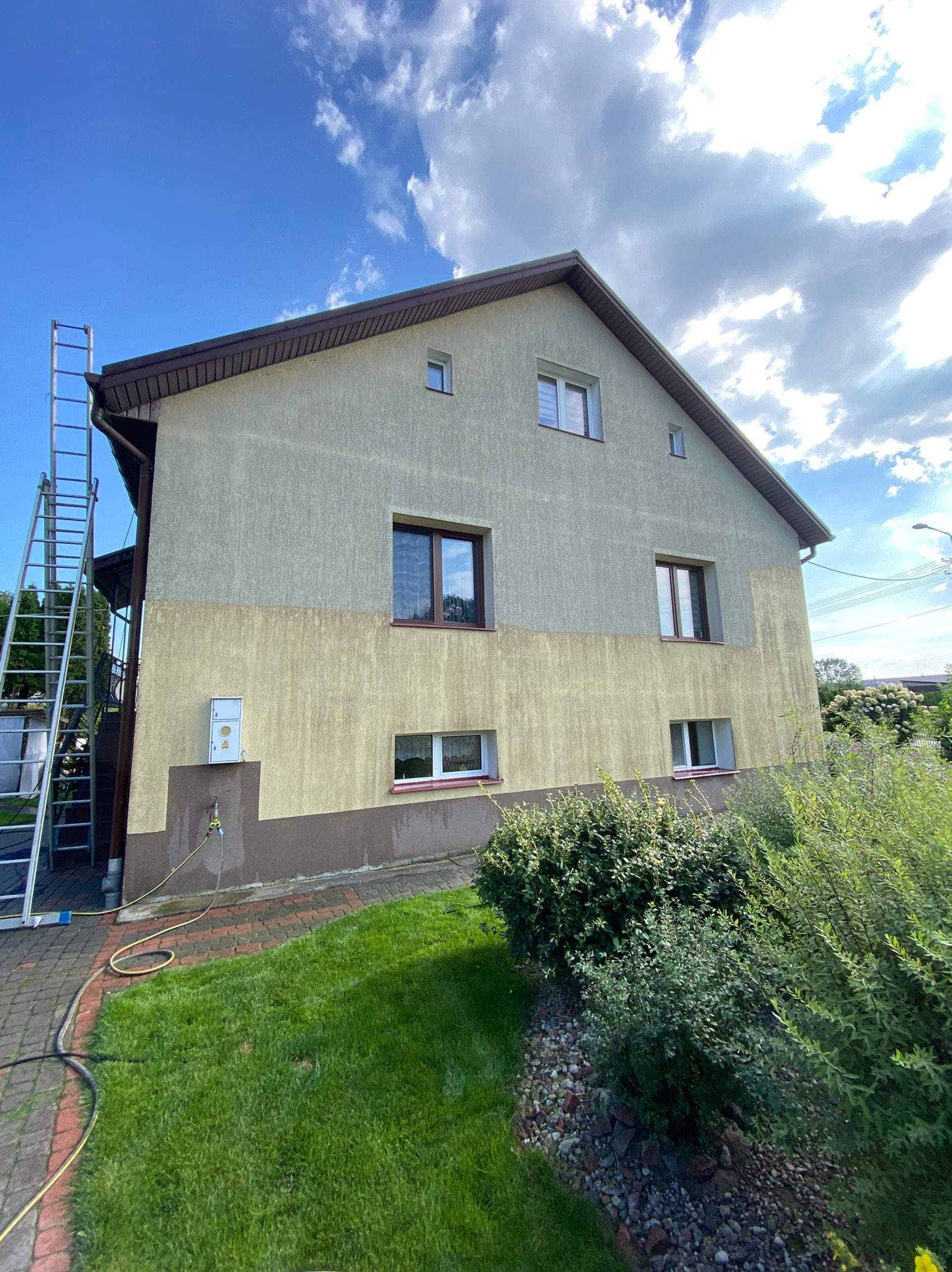 Dwukondygnacyjny dom z elewacją w trakcie mycia, widoczna drabina i wąż ogrodowy. Część ściany jest czysta, a część brudna. Słoneczny dzień, błękitne niebo.