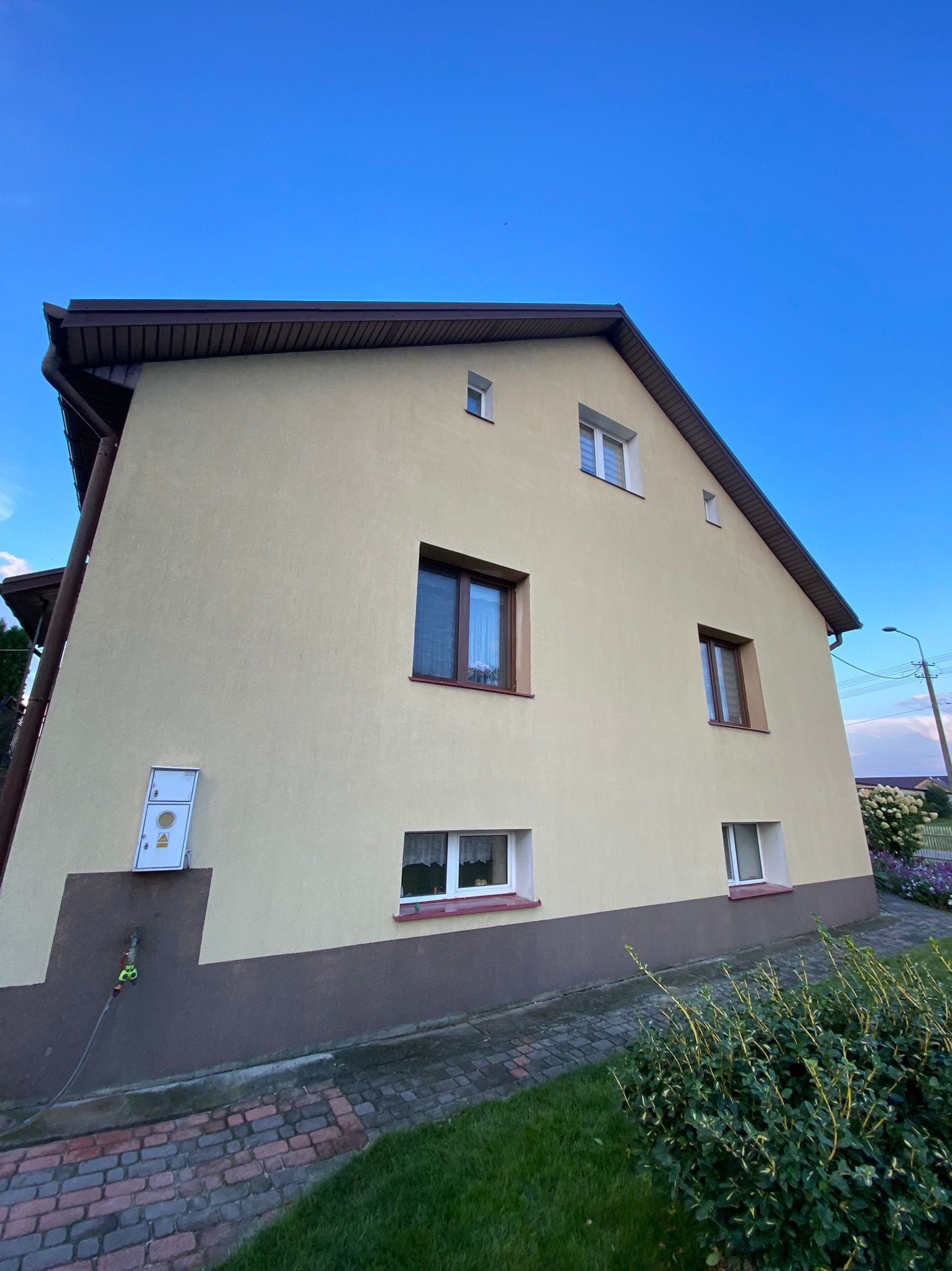 Dwukondygnacyjny dom z beżową elewacją i brązowym cokołem, widziany pod kątem z dołu, na tle błękitnego nieba. Widoczny fragment ogrodu i skrzynka elektryczna na ścianie.
