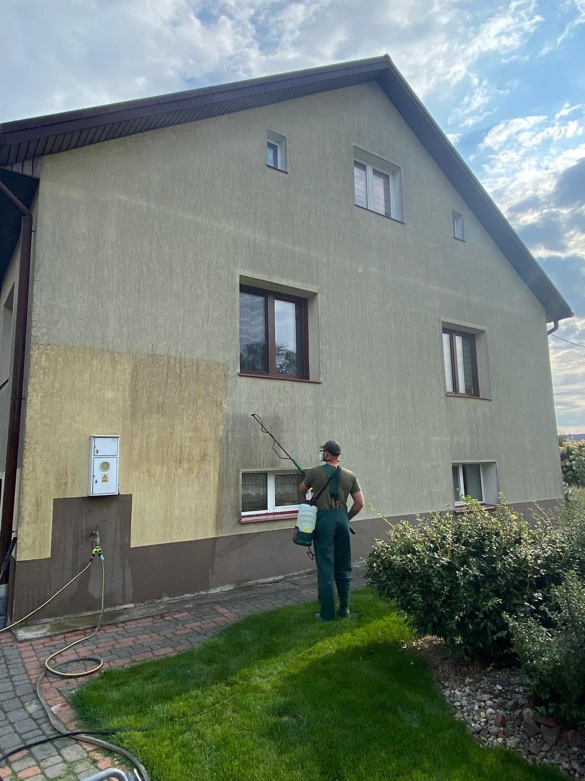 Mężczyzna w kombinezonie spryskuje ścianę domu, widoczny efekt czyszczenia elewacji. Część ściany jest już umyta, a część jeszcze zabrudzona.
