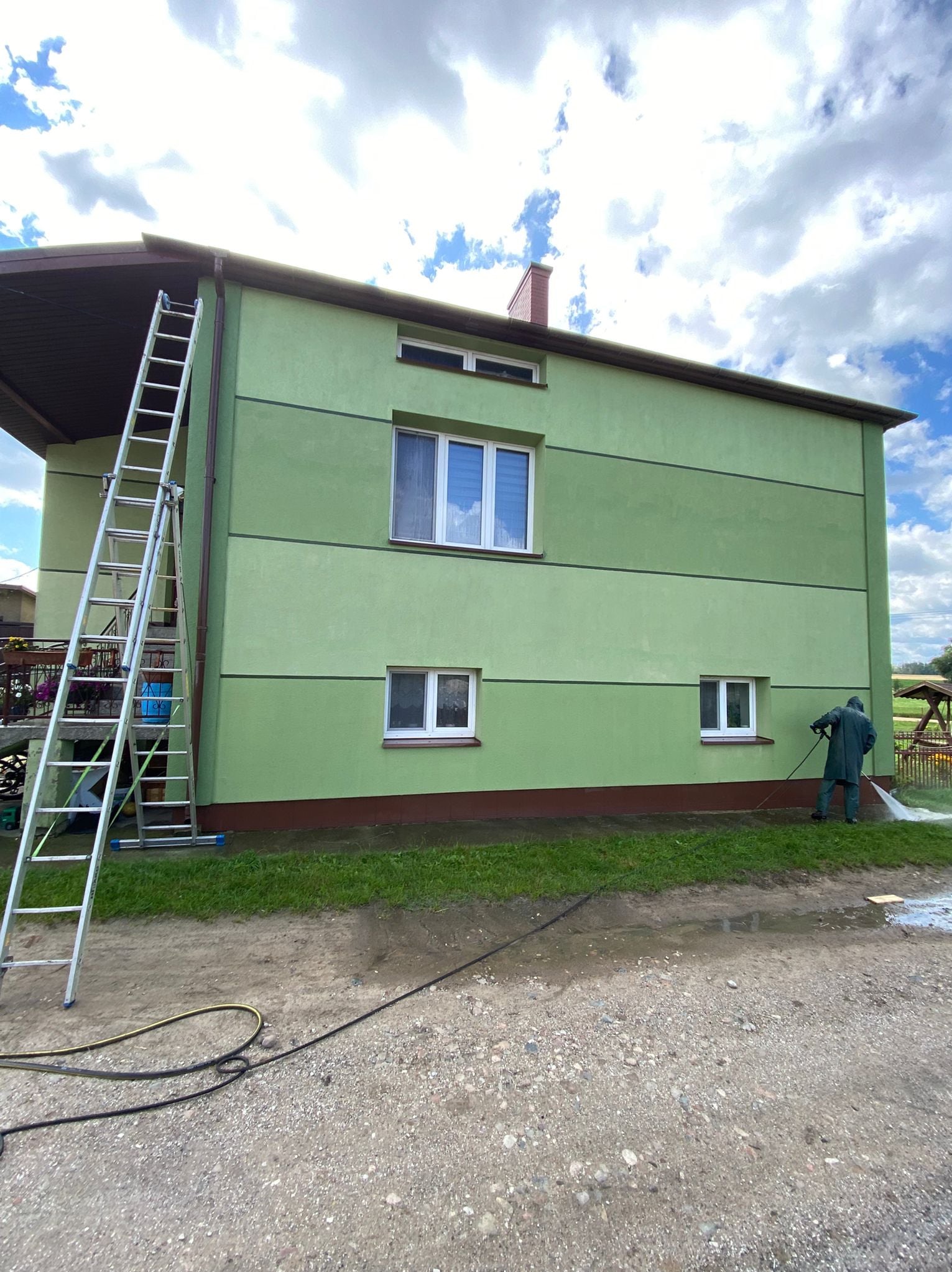 Zielona elewacja domu po myciu ciśnieniowym. Widoczna drabina oparta o ścianę i osoba w kombinezonie czyszcząca elewację. Błękitne niebo z chmurami w tle.