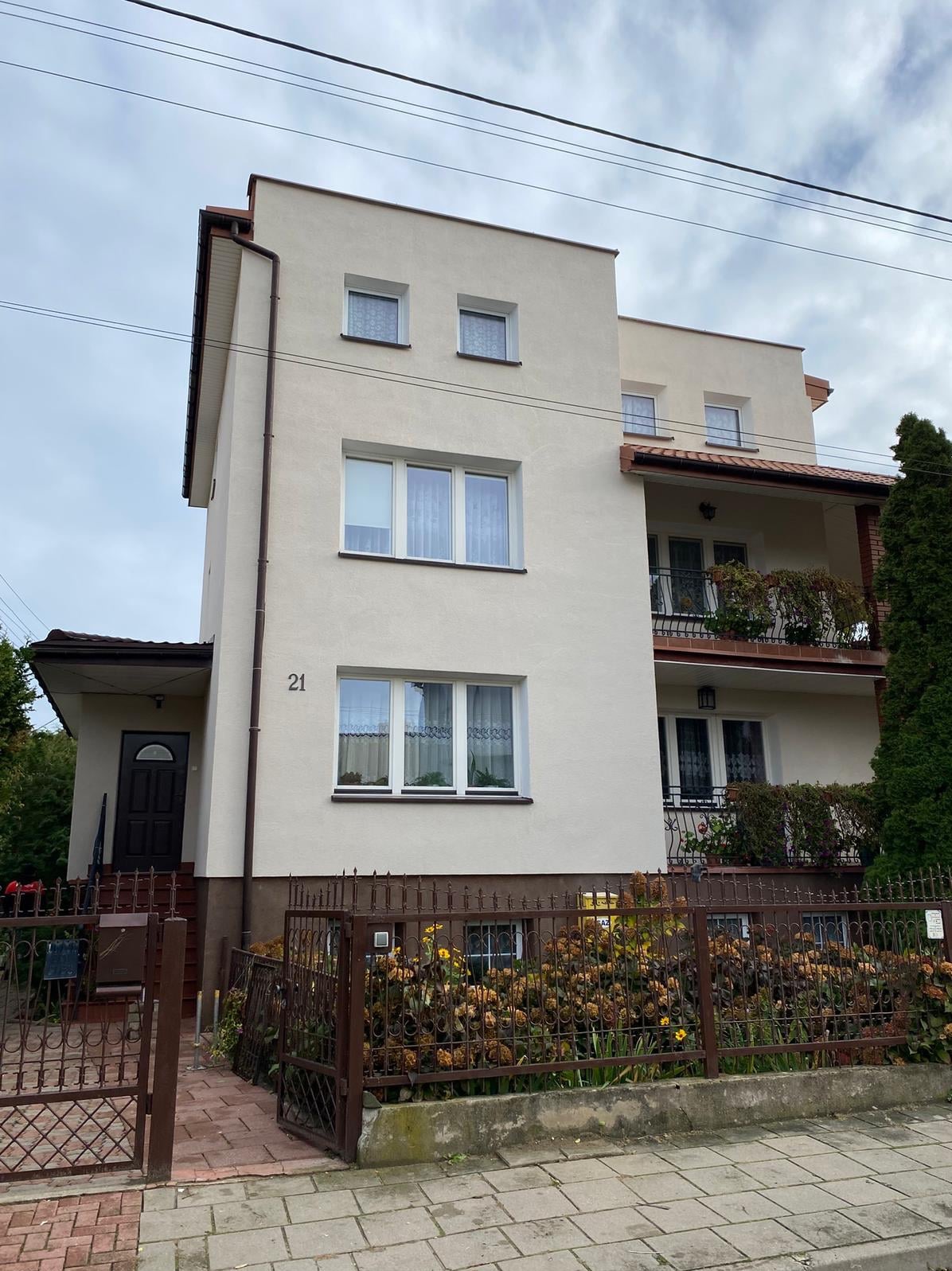 Trzykondygnacyjny dom z beżową elewacją, balkonami z kutymi balustradami i numerem '21' na ścianie, widziany zza metalowego ogrodzenia.