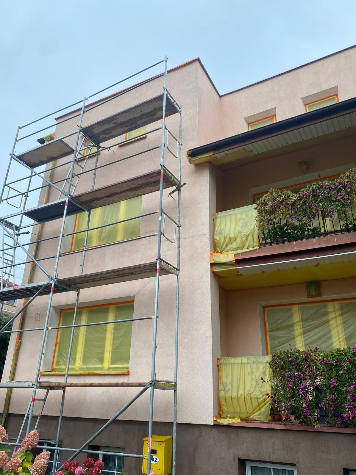 Budynek w trakcie zabezpieczania elewacji folią i taśmą malarską przed pracami. Widoczne rusztowania i kwiaty na balkonie. Ujęcie z dołu.