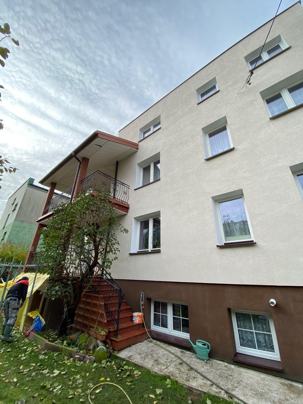 Odnowiona elewacja budynku po malowaniu, z widocznymi oknami i balkonem. Na pierwszym planie pracownik w trakcie sprzątania, zieleń i narzędzia ogrodowe.