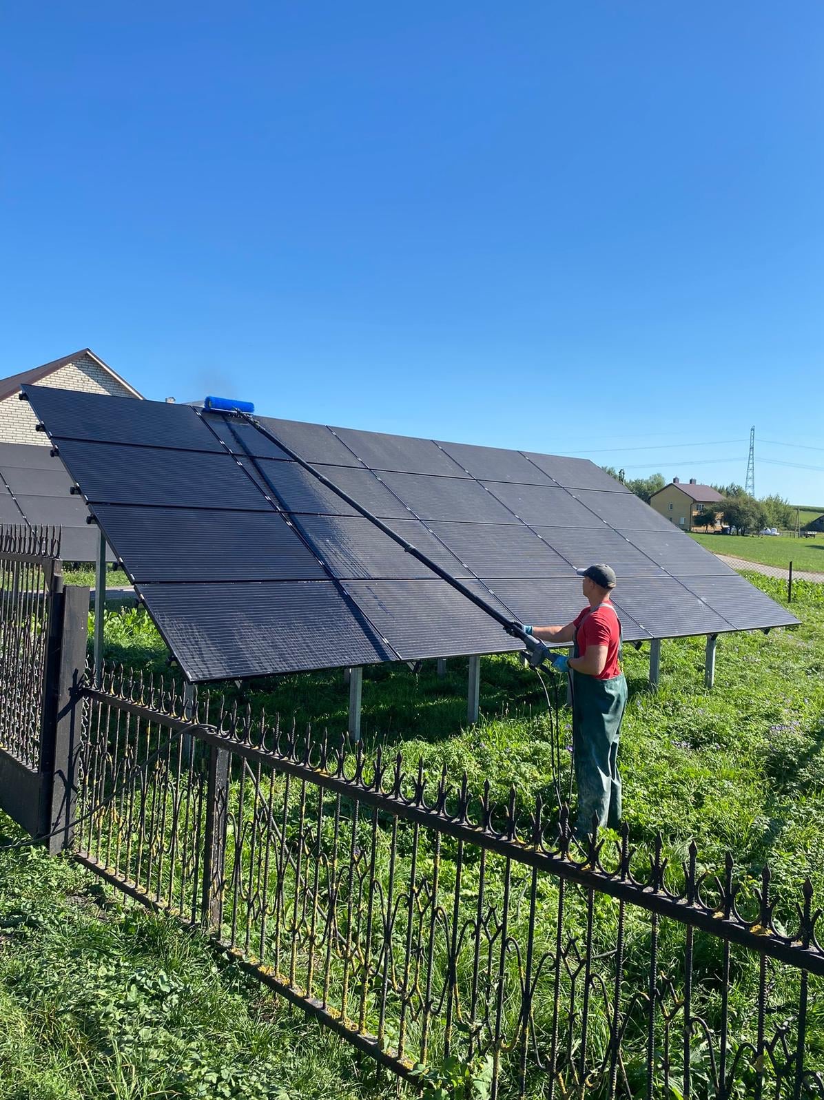 Czyszczenie paneli fotowoltaicznych na trawniku, pracownik w kombinezonie używa szczotki na wysięgniku. W tle dom i błękitne niebo.