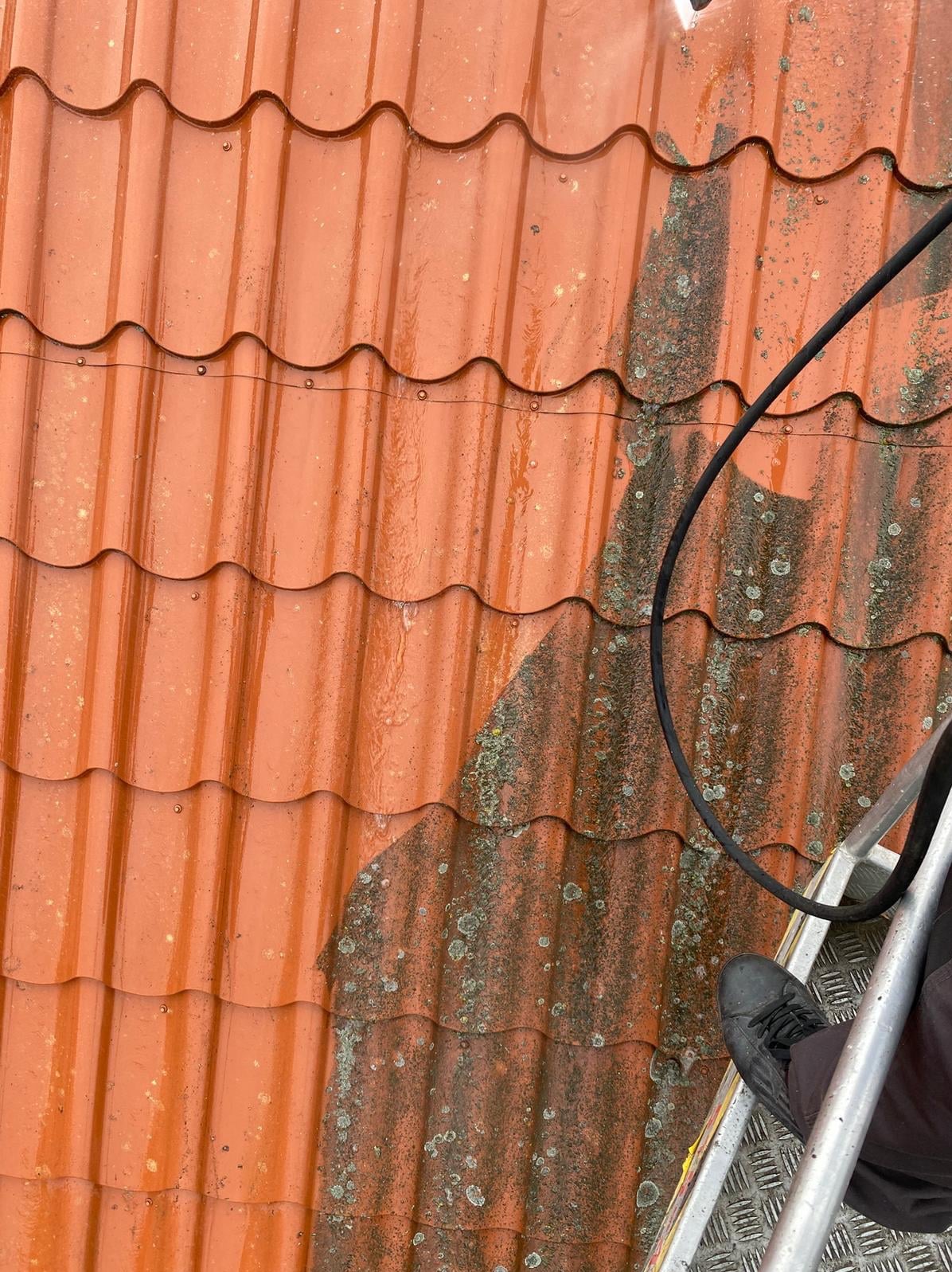 Częściowo umyty dach z czerwonej dachówki, widoczne zabrudzenia i mech. W tle wąż ciśnieniowy i fragment drabiny. Ukazany efekt czyszczenia.
