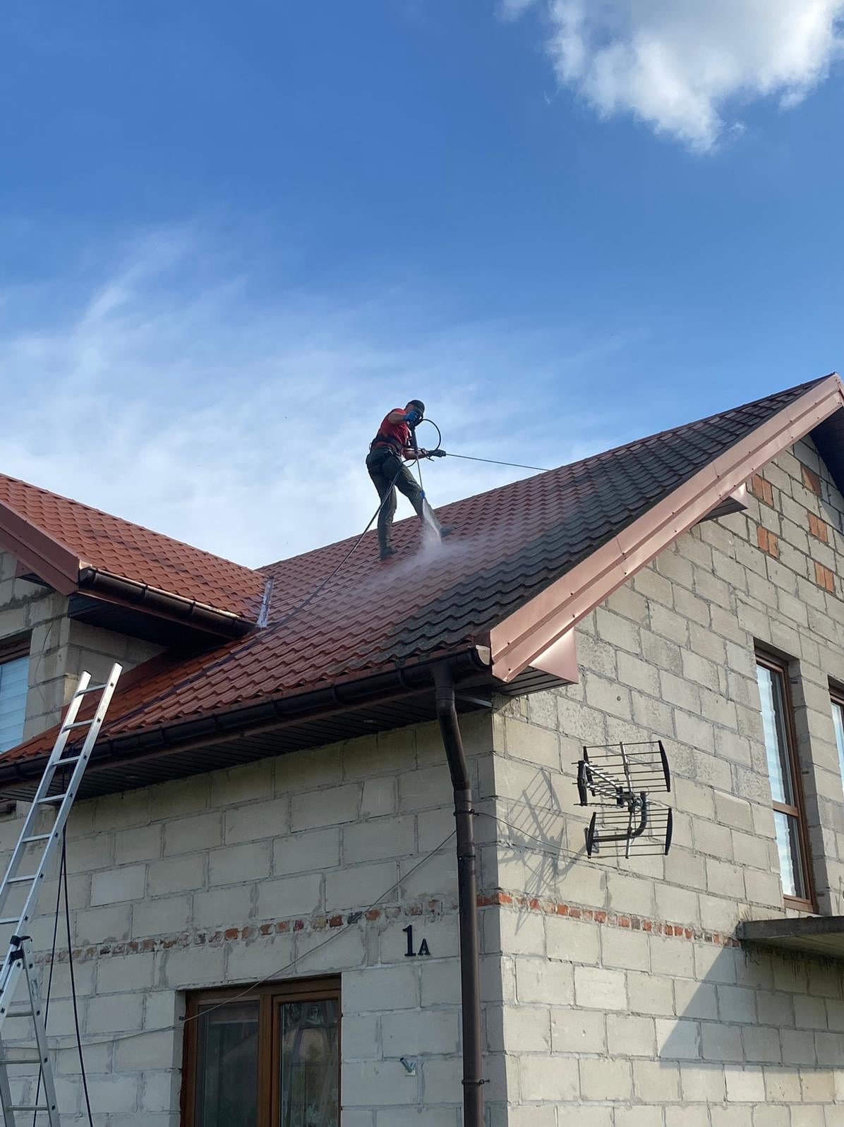 Pracownik w kasku i masce czyści dach z brązowej dachówki za pomocą myjki ciśnieniowej. Widoczna drabina oparta o budynek z szarego pustaka i błękitne niebo.