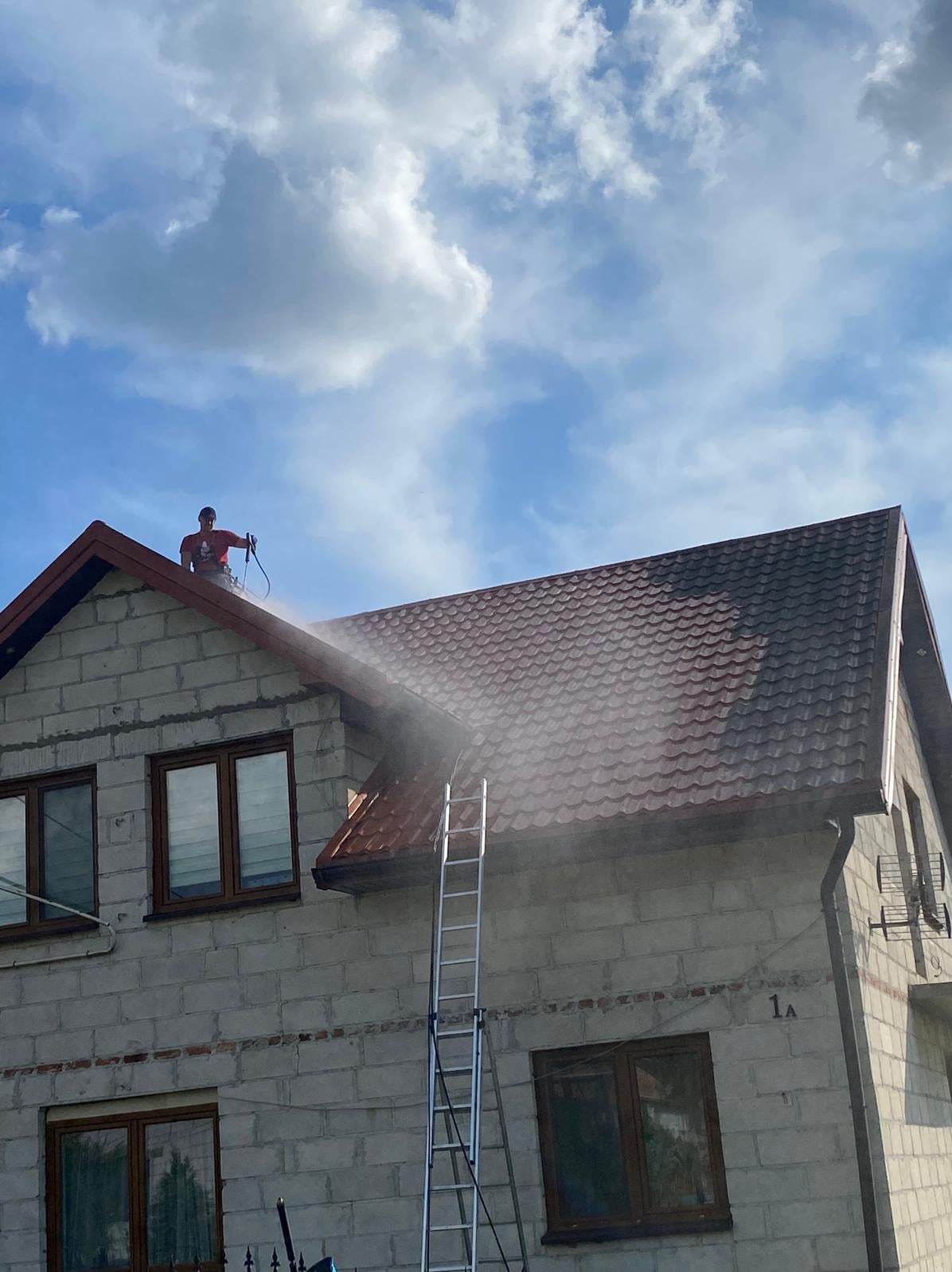 Czyszczenie dachu z drabiny przez pracownika w czerwonej koszulce, strumień wody pod ciśnieniem, błękitne niebo z chmurami w tle. Dachówka w kolorze brązowym.