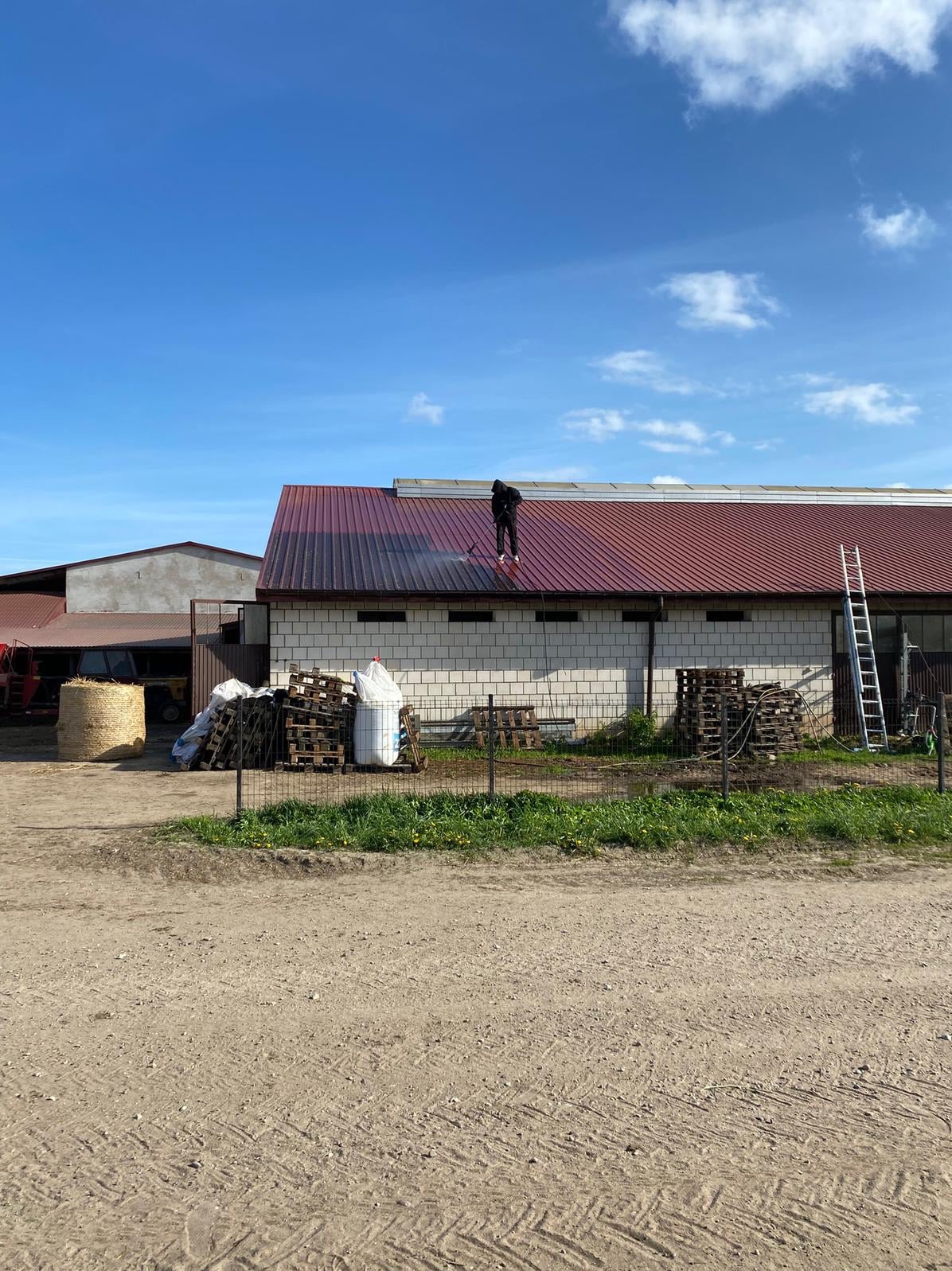 Pracownik w trakcie mycia dachu budynku gospodarczego z czerwonej blachy, widoczna drabina i stos palet. Błękitne niebo w tle.