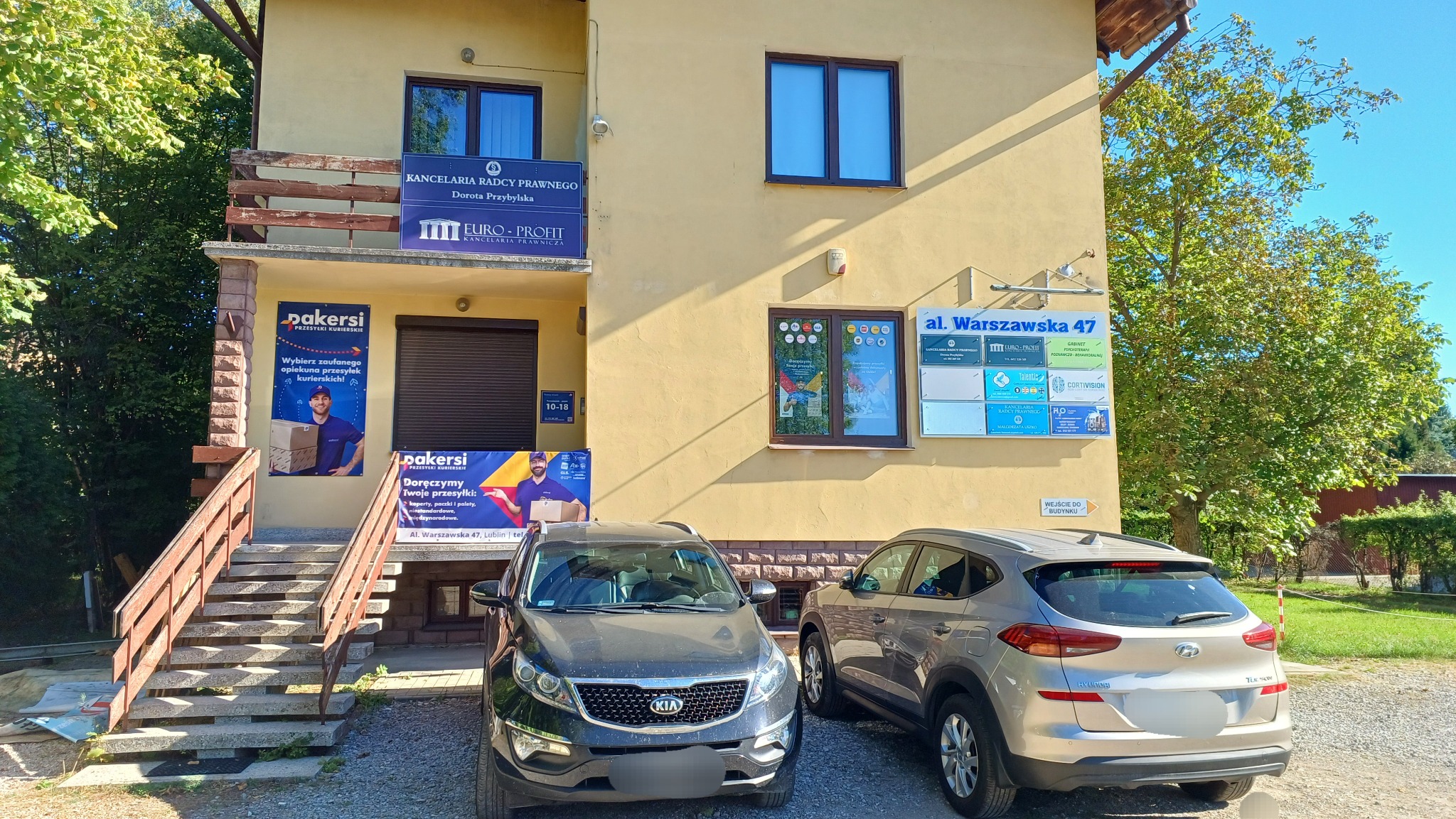 Budynek firmy kurierskiej Dakersi z samochodami na parkingu, reklamy na elewacji i tablica z adresem Al. Warszawska 47 w Lublinie. Słoneczny dzień.