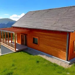 Drewniany dom z gontowym dachem na tle gór i błękitnego nieba. Widoczny taras z drewnianą konstrukcją i zielony trawnik. Ujęcie z lotu ptaka.