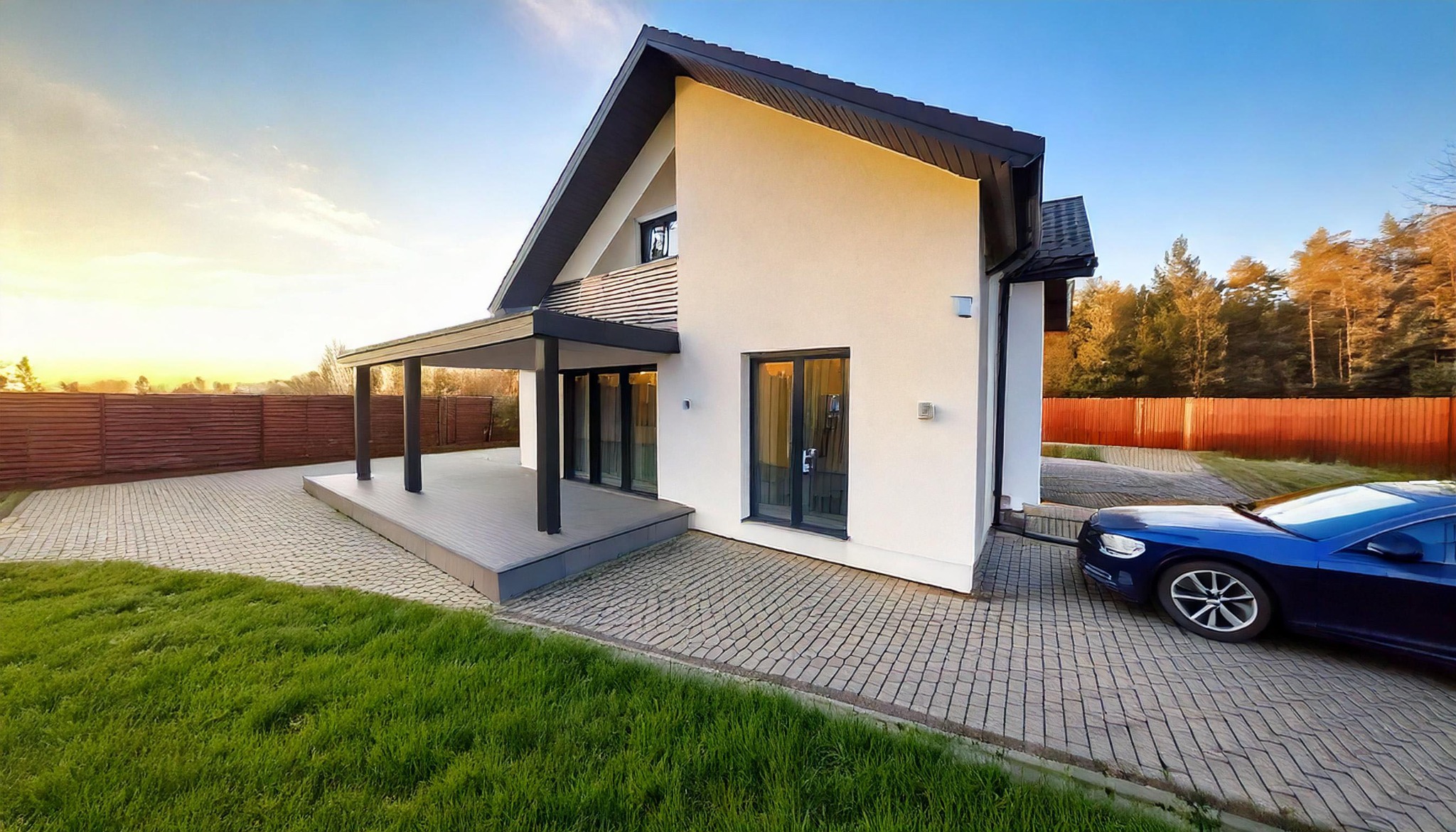 Elegancki, nowoczesny dom jednorodzinny z tarasem i podjazdem z kostki, na tle zielonego trawnika i drzew. Błękitny samochód zaparkowany obok budynku.