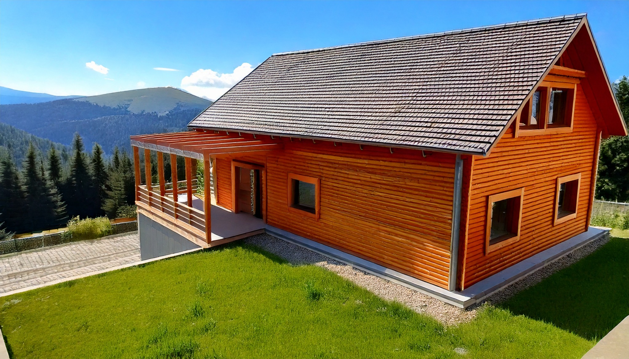 Drewniany dom z gontowym dachem na tle gór i błękitnego nieba. Widoczny taras z drewnianą konstrukcją i zielony trawnik. Ujęcie z lotu ptaka.
