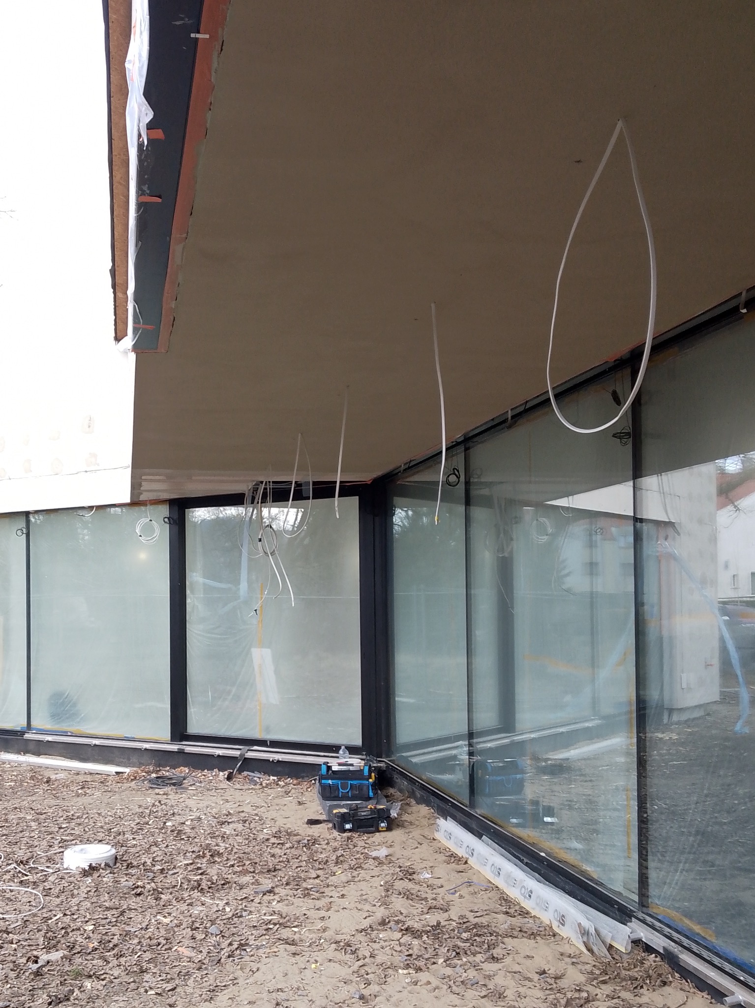 Wnętrze budynku w trakcie prac wykończeniowych; widoczne kable zwisające z sufitu, okna z folią ochronną i narzędzia elektryczne na podłodze.