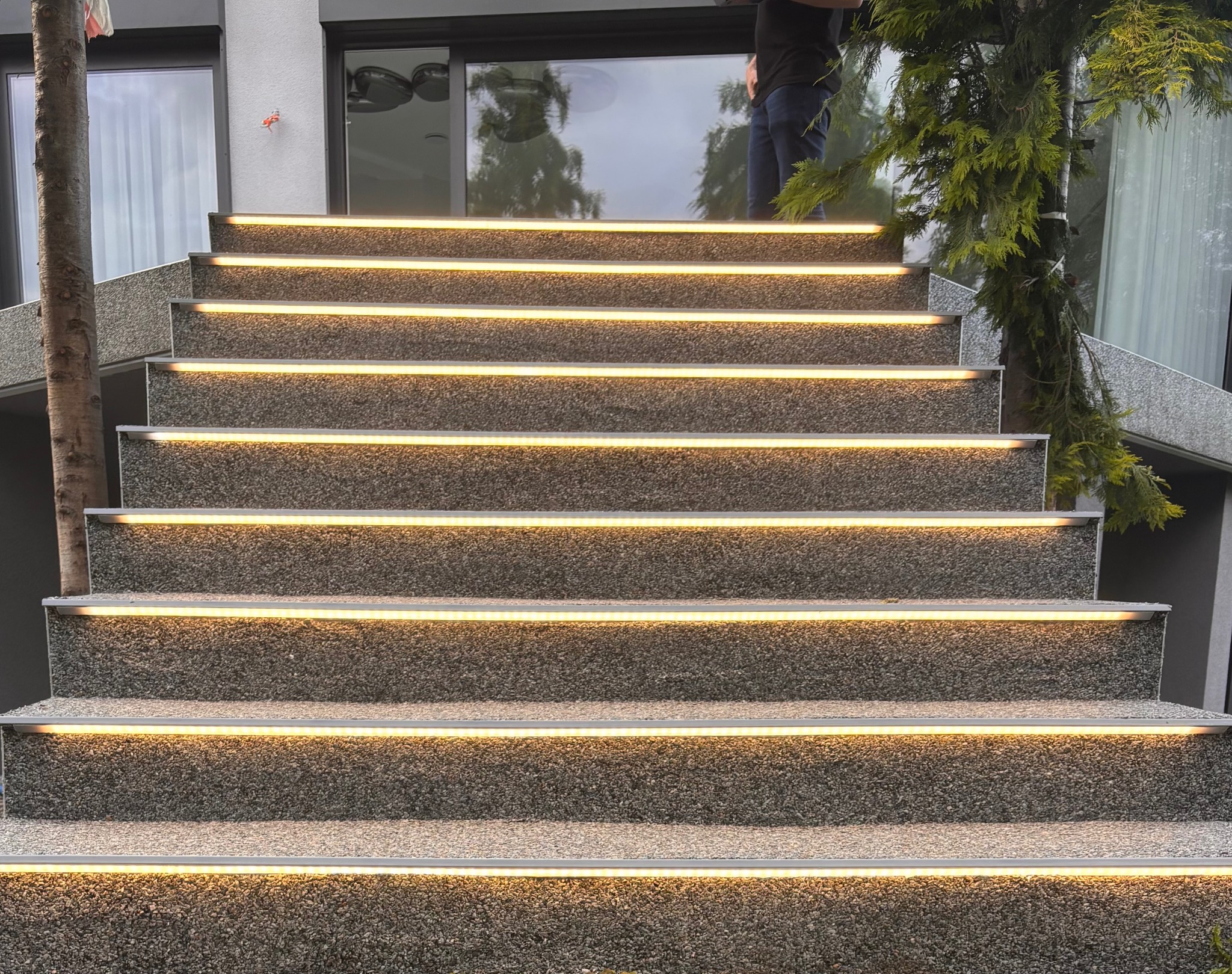 Schody zewnętrzne z oświetleniem LED wzdłuż krawędzi stopni, wykonane z szarego kamienia, z widocznym fragmentem osoby na górze i elementami zieleni po bokach.