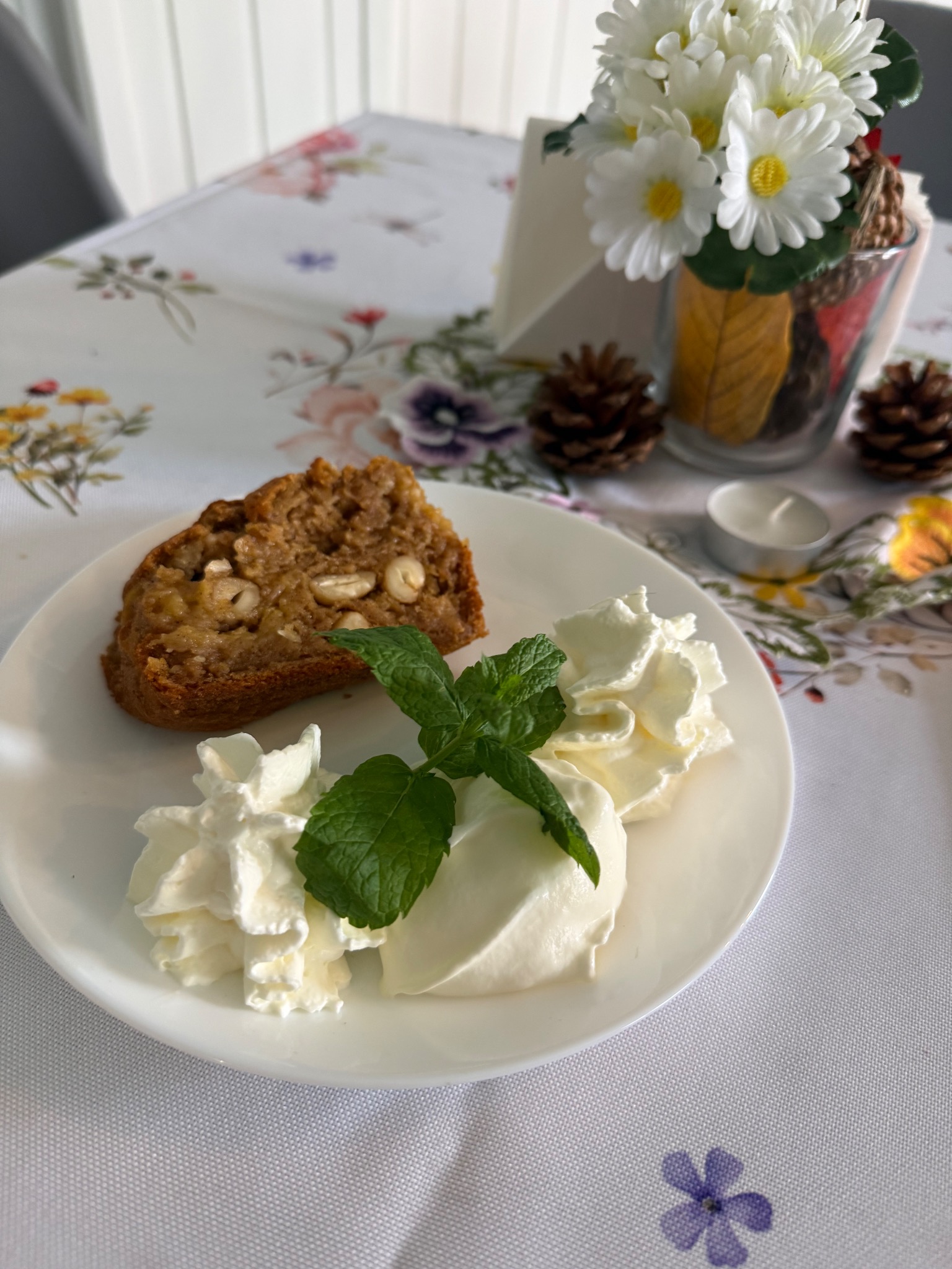 Kawałek ciasta orzechowego z bitą śmietaną i miętą na białym talerzu, dekoracja stołu z kwiatami i szyszkami w tle.