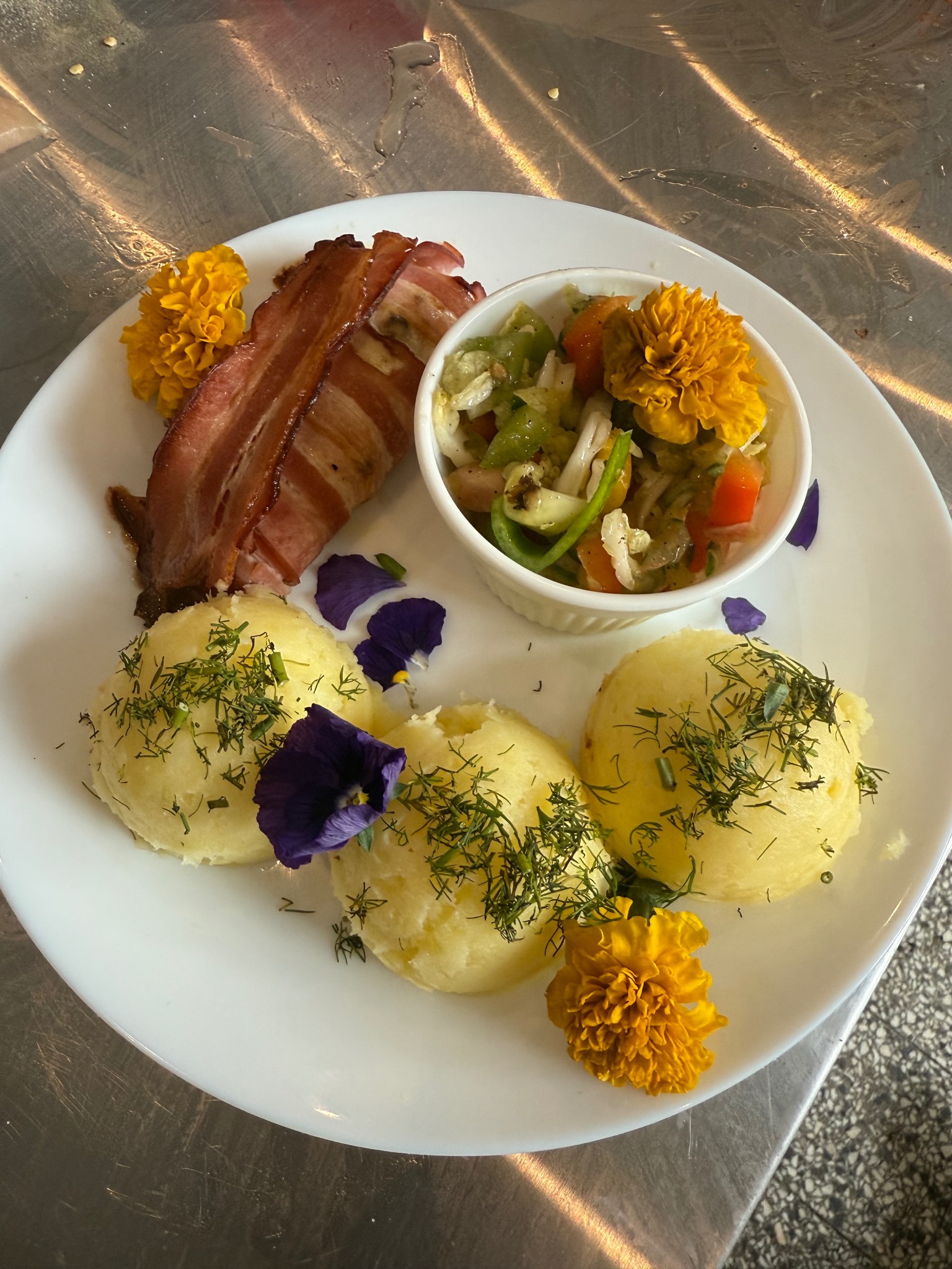 Elegancko podane danie: ziemniaki z koperkiem, sałatka warzywna w białej miseczce i grillowany boczek, ozdobione kwiatami na białym talerzu.