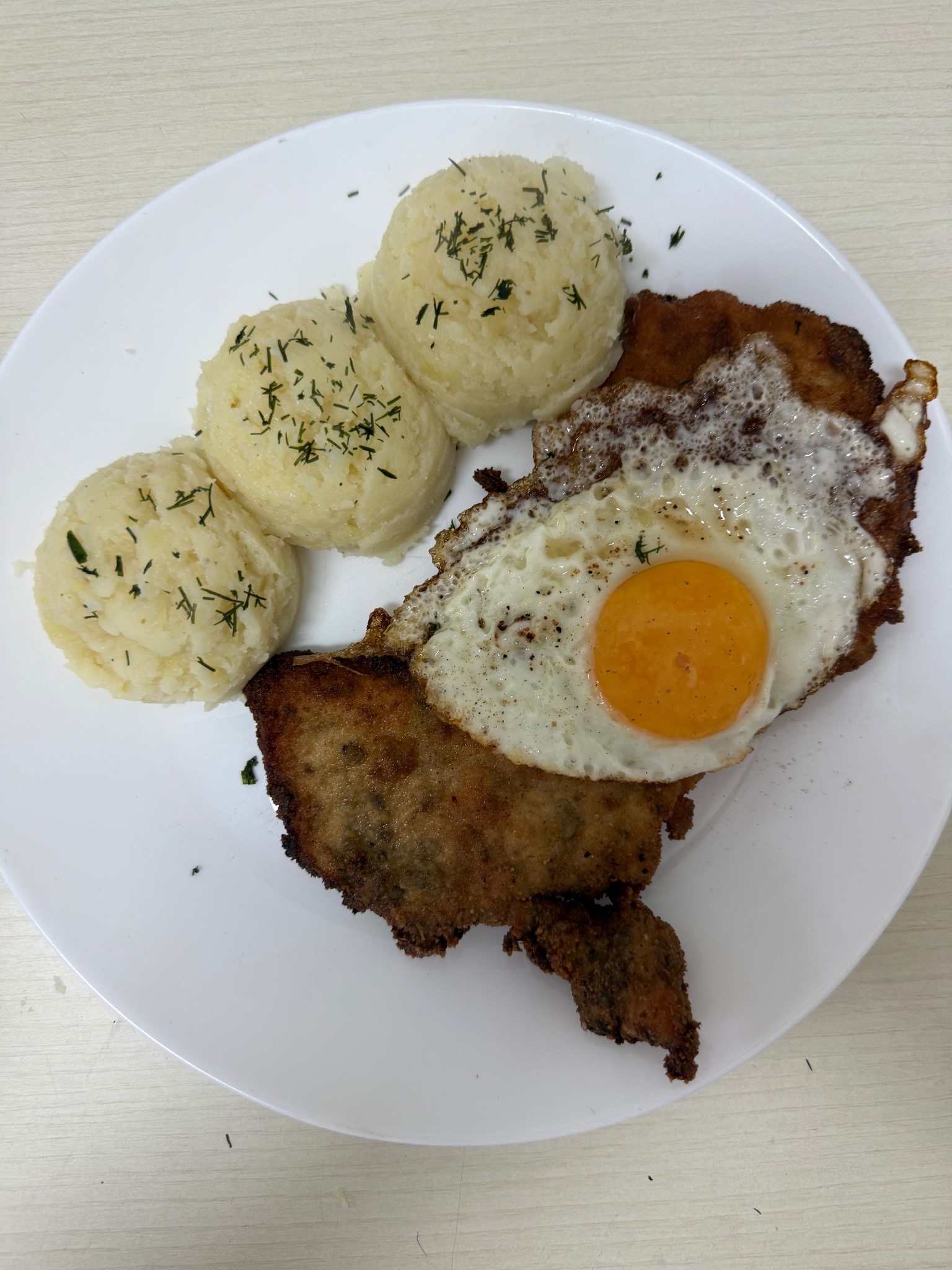 Kotlet schabowy z jajkiem sadzonym i trzema porcjami ziemniaków puree posypanych koperkiem na białym talerzu, całość sfotografowana z góry.