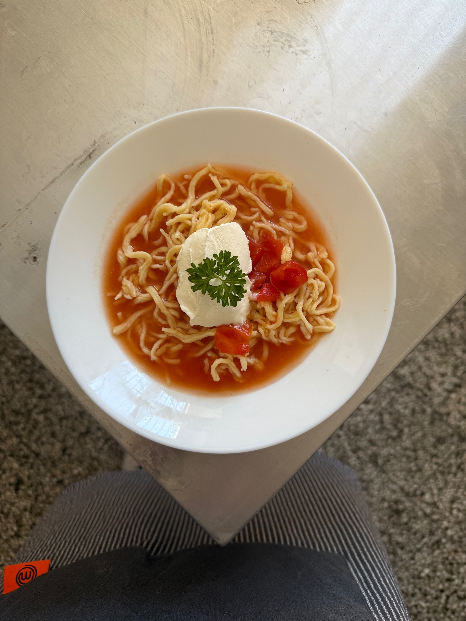 Talerz z zupą pomidorową z makaronem, kleksem śmietany i posiekanymi pomidorami, udekorowany zieloną pietruszką. Zupa podana na metalowym stole.