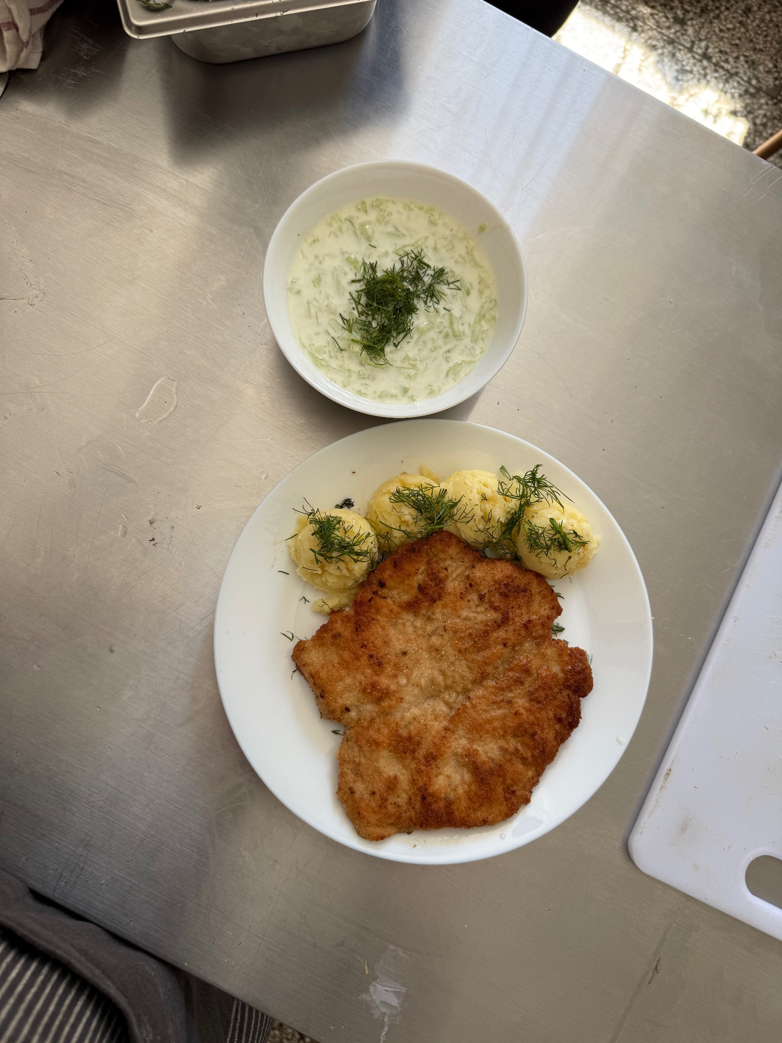 Kotlet schabowy z ziemniakami i mizerią, udekorowany koperkiem, na białym talerzu i w miseczce, na metalowym blacie. Widok z góry.