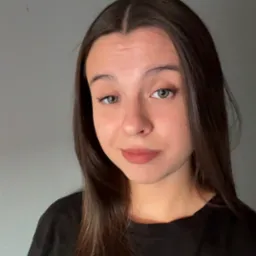 Portret młodej kobiety z długimi, ciemnymi włosami i zielonymi oczami, ubrana w czarną koszulkę, na jednolitym, szarym tle. Uśmiech delikatny, skierowany wprost do widza.