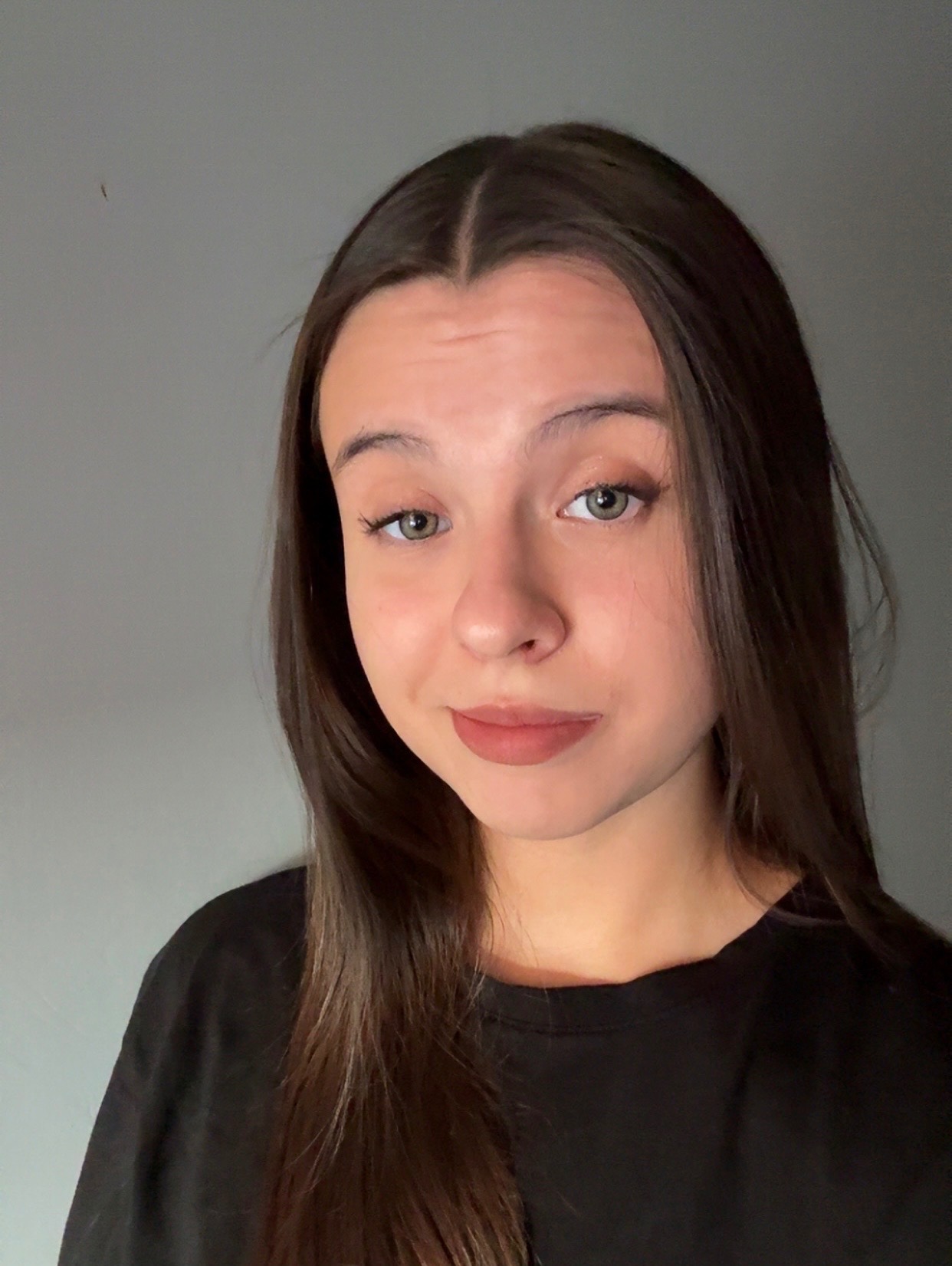 Portret młodej kobiety z długimi, ciemnymi włosami i zielonymi oczami, ubrana w czarną koszulkę, na jednolitym, szarym tle. Uśmiech delikatny, skierowany wprost do widza.