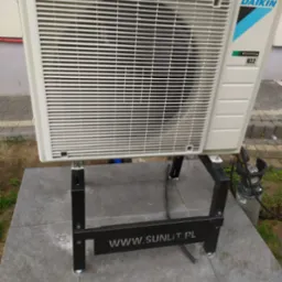 Biała jednostka zewnętrzna klimatyzacji Daikin na czarnym stelażu z widocznym adresem www.sunlit.pl, ustawiona na szarych płytach chodnikowych.
