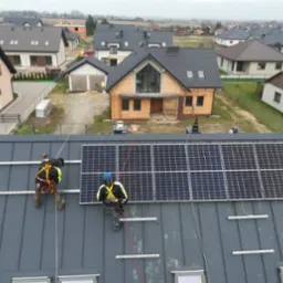 Montaż paneli fotowoltaicznych na szarym dachu domu jednorodzinnego przez dwóch pracowników w uprzężach, z widokiem na okoliczne budynki.