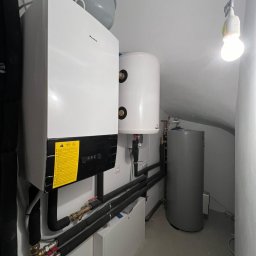 Sunlit - Biały piec grzewczy Daikin z żółtą naklejką, zbiornik wyrównawczy i dwa zasobniki ciepłej wody w kotłowni. Widoczne rury i zawory.