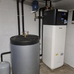 Sunlit - Zestaw grzewczy Daikin z zbiornikiem i rurami w kotłowni wyłożonej białymi płytkami. Widoczne manometry i zawory, instalacja pod sufitem.