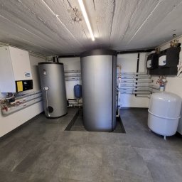 Sunlit - Kotłownia z dwoma srebrnymi zbiornikami akumulacyjnymi, białym piecem i instalacją rurową z zaworami na ścianach. Podłoga wyłożona szarymi płytkami.