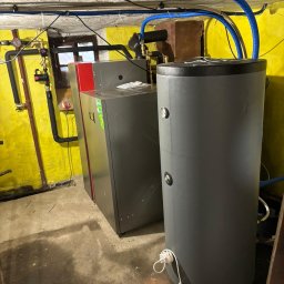 Sunlit - Szary piec CO z czerwonym panelem bocznym i bojler w piwnicy z żółtymi ścianami, widoczne rury instalacyjne i okno.