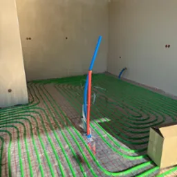 Zielone rurki ogrzewania podłogowego ułożone na czerwonej siatce w surowym wnętrzu. Pionowe rury niebieskie i czerwone wystają z podłoża. Widoczny karton.