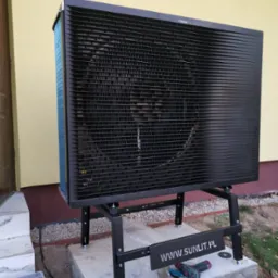 Czarna pompa ciepła Daikin na betonowym fundamencie, z narzędziami i tabliczką www.sunlit.pl. Żółta ściana budynku w tle. Widok z boku, częściowo z dołu.