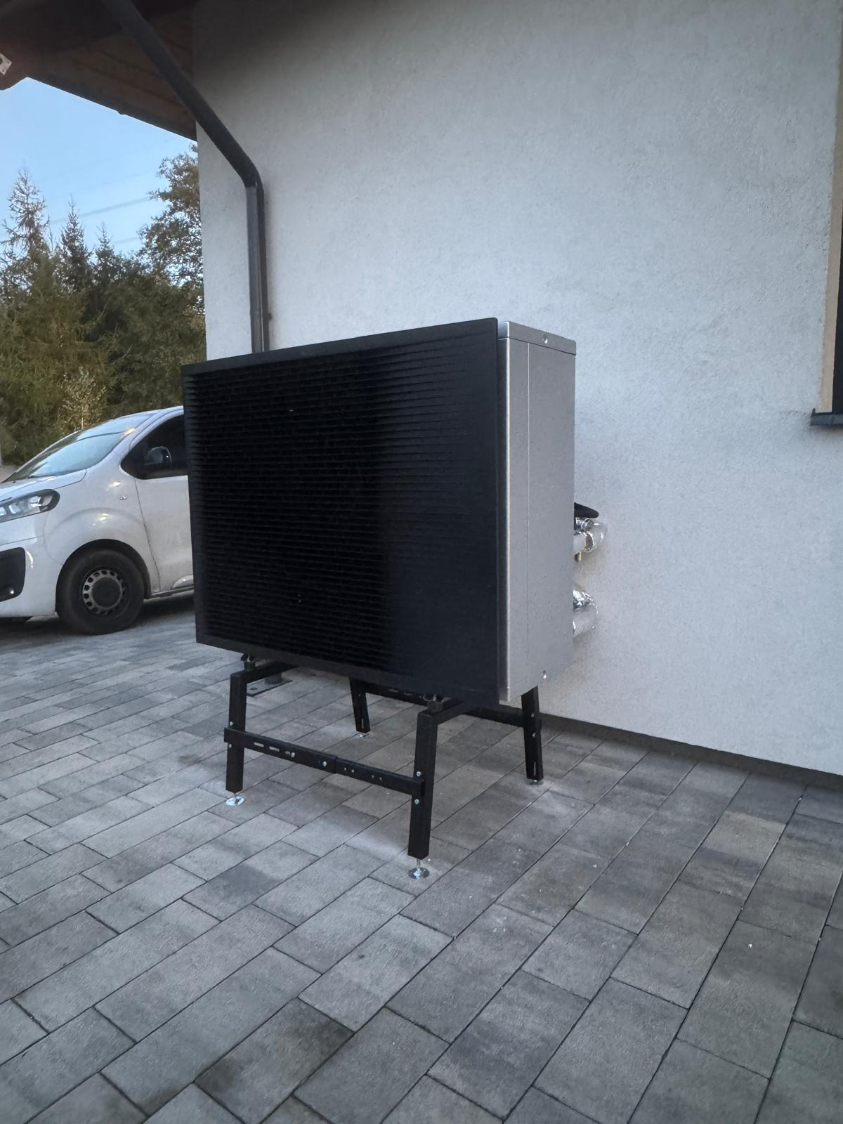 Zewnętrzna jednostka pompy ciepła na metalowej konstrukcji, ustawiona na kostce brukowej przy ścianie domu. Widoczny fragment białego samochodu.