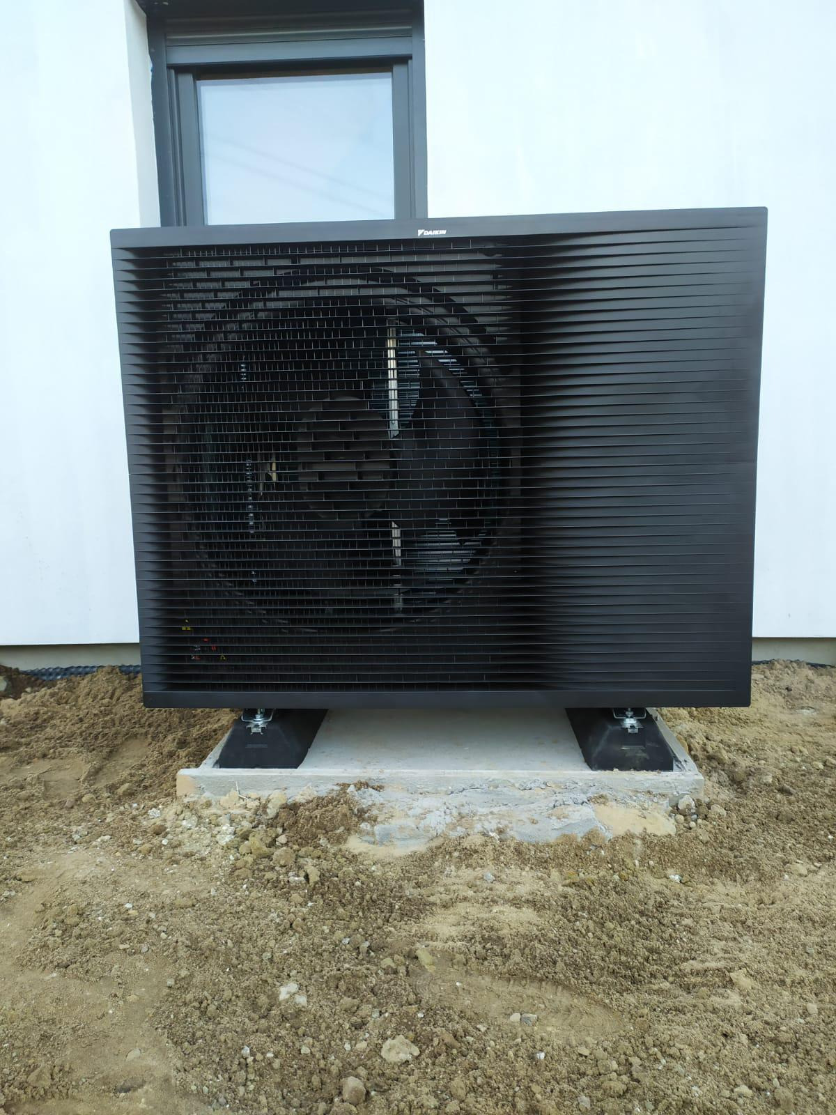Czarna pompa ciepła Daikin na betonowej podstawie, ustawiona przy białej ścianie budynku z oknem. Widoczny fragment terenu z ziemią.